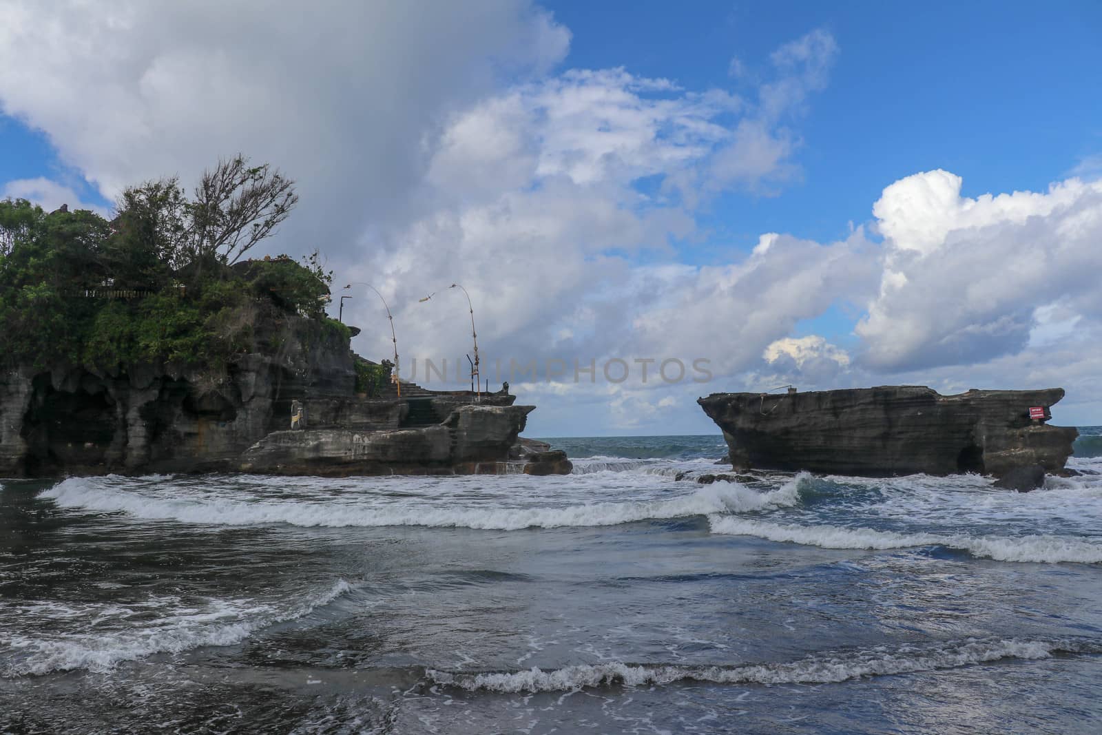 Waves shatter on a cliff at the top of which is the Hindu temple of Tanah Lot. Temple built on a rock in the sea off the coast of Bali island, Indonesia. Lush tropical vegetation on top of a cliff.
