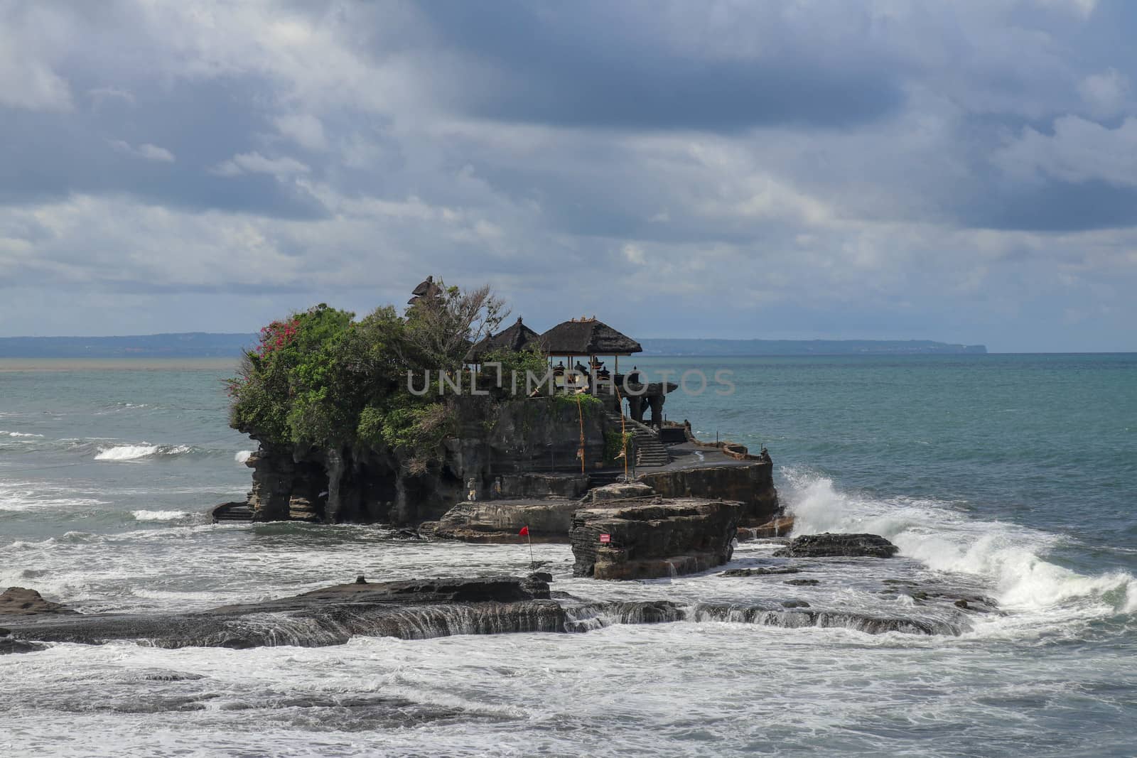 Aerial view of Tanah Lot temple on cliff in Indian ocean. Waves are breaking on a rock cliff off the coast of Bali. Hindu temple built on a rock in the sea. Lush tropical vegetation on top of a cliff.