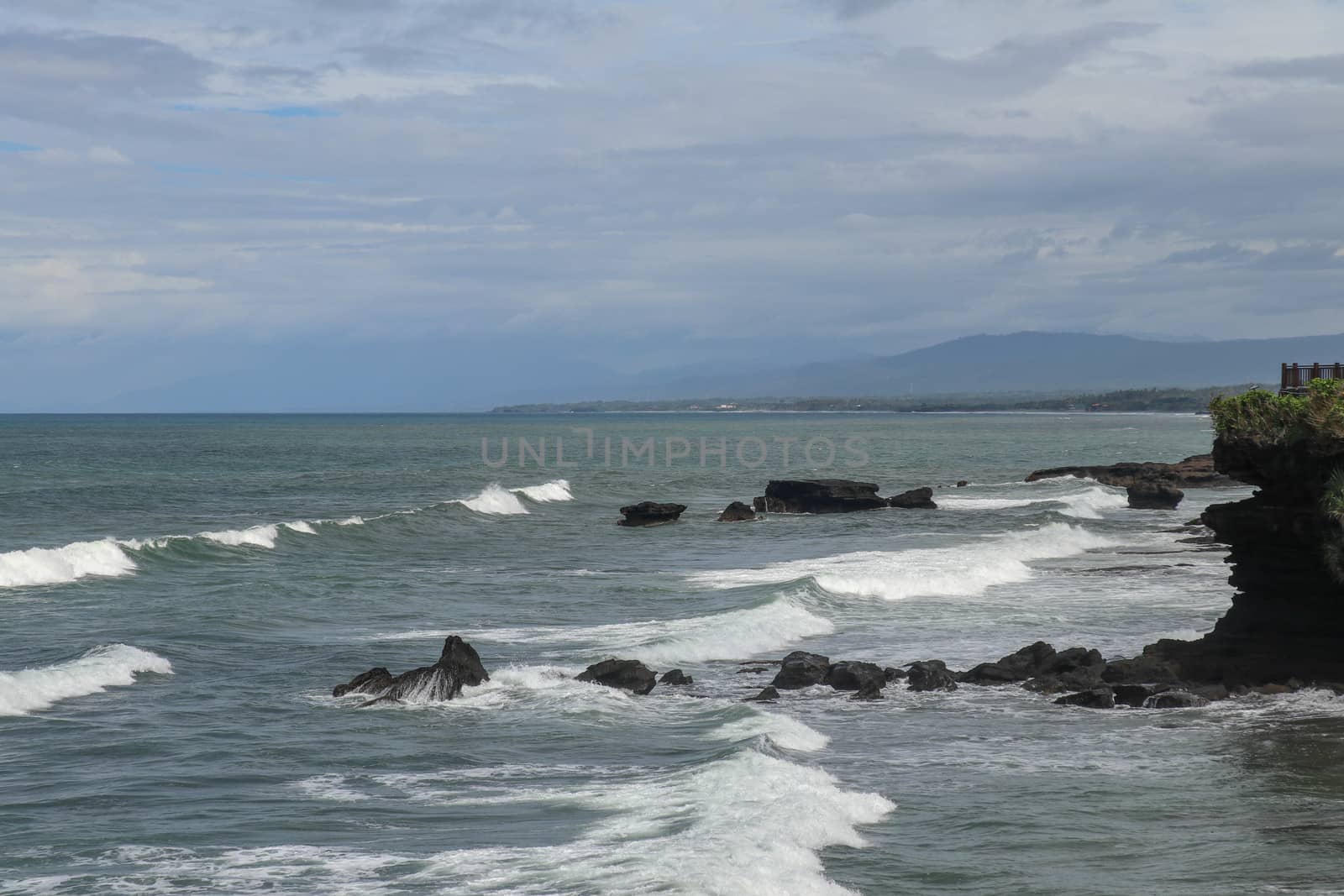 Rock cliffs in the Indian Ocean off the coast of Bali island, near Batu Bolong and tourism attraction spot Tanah Lot temple. Waves are shattering on rocks of solidified black volcanic lava. Indonesia.