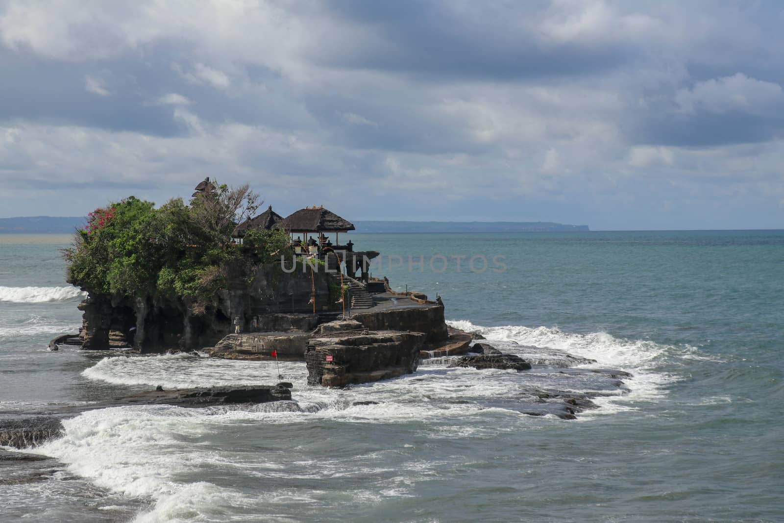Aerial view of Tanah Lot temple on cliff in Indian ocean. Waves are breaking on a rock cliff off the coast of Bali. Hindu temple built on a rock in the sea. Lush tropical vegetation on top of a cliff.