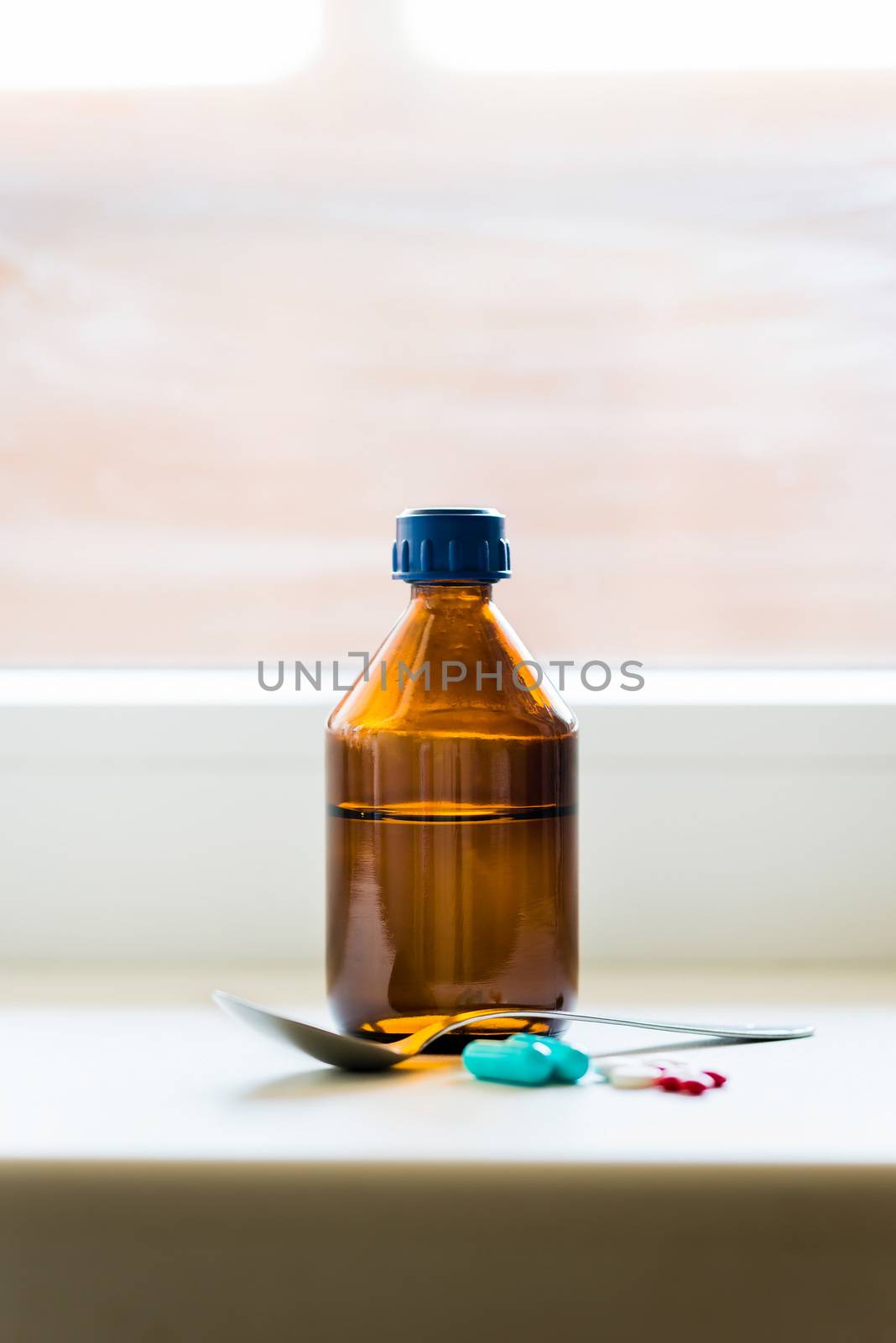 A cough syrup bottle with a spoon and some pills close to the window