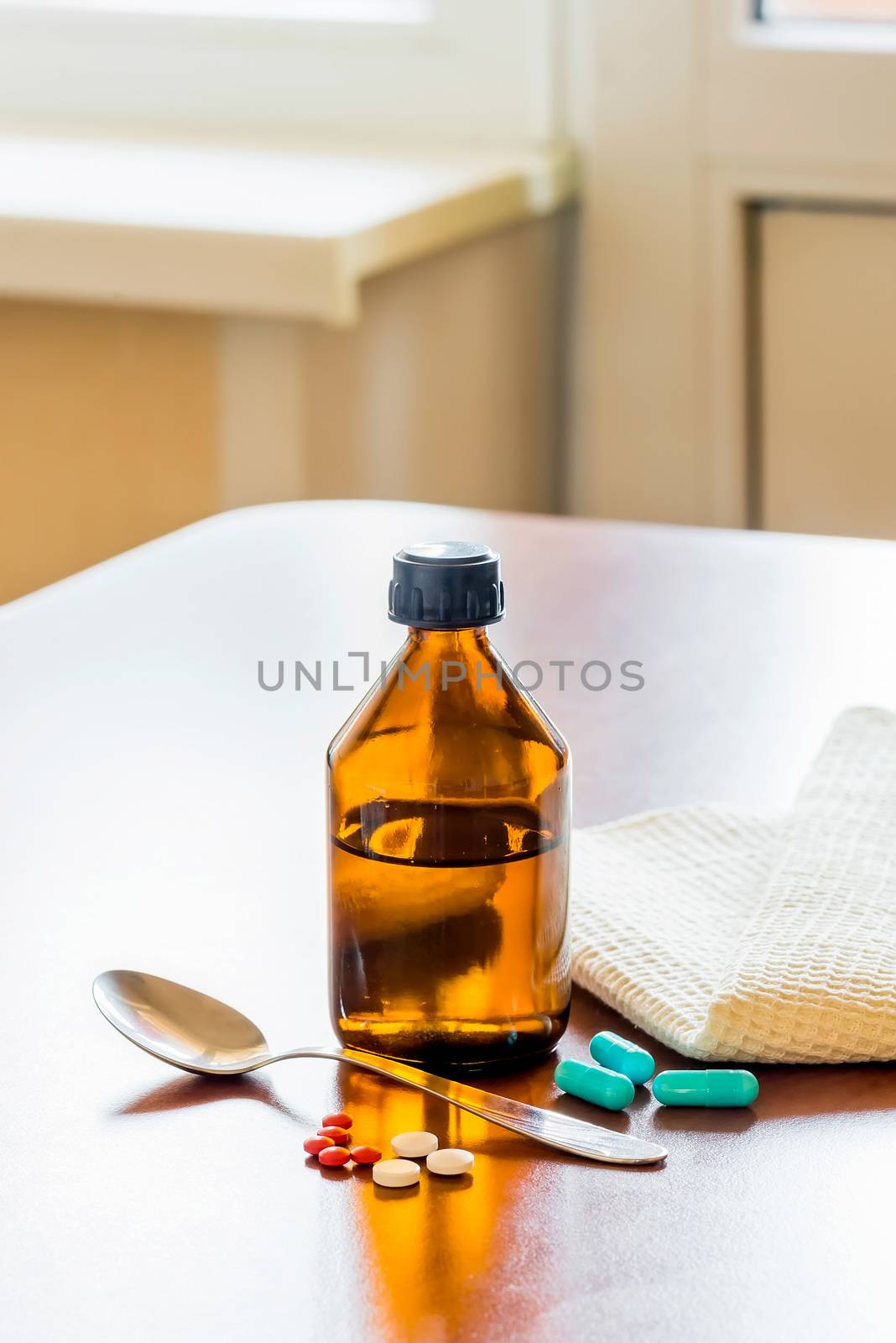 A cough syrup bottle with a spoon and some pills on the table close to the window
