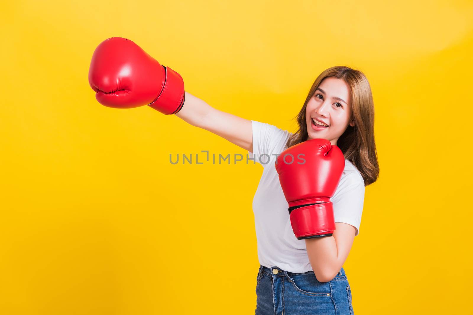 Portrait Asian Thai beautiful young woman standing smile wearing red boxing gloves and punch to side away space, studio shot isolated on yellow background, There was copy space for text