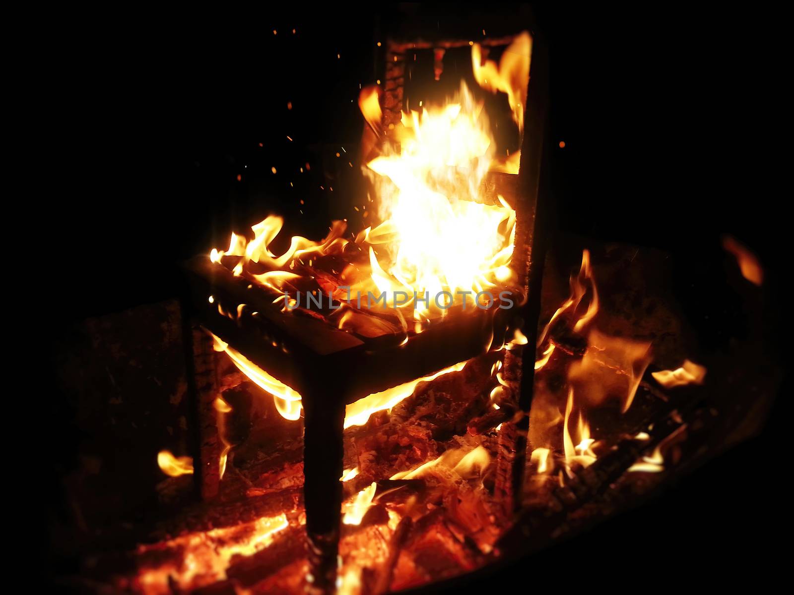 Wooden chair burning inside the bonfire. Close up on sparks rises from the fire with heatwaves at night. A horror concept of a demonic ritual or devil and demons religious worship.