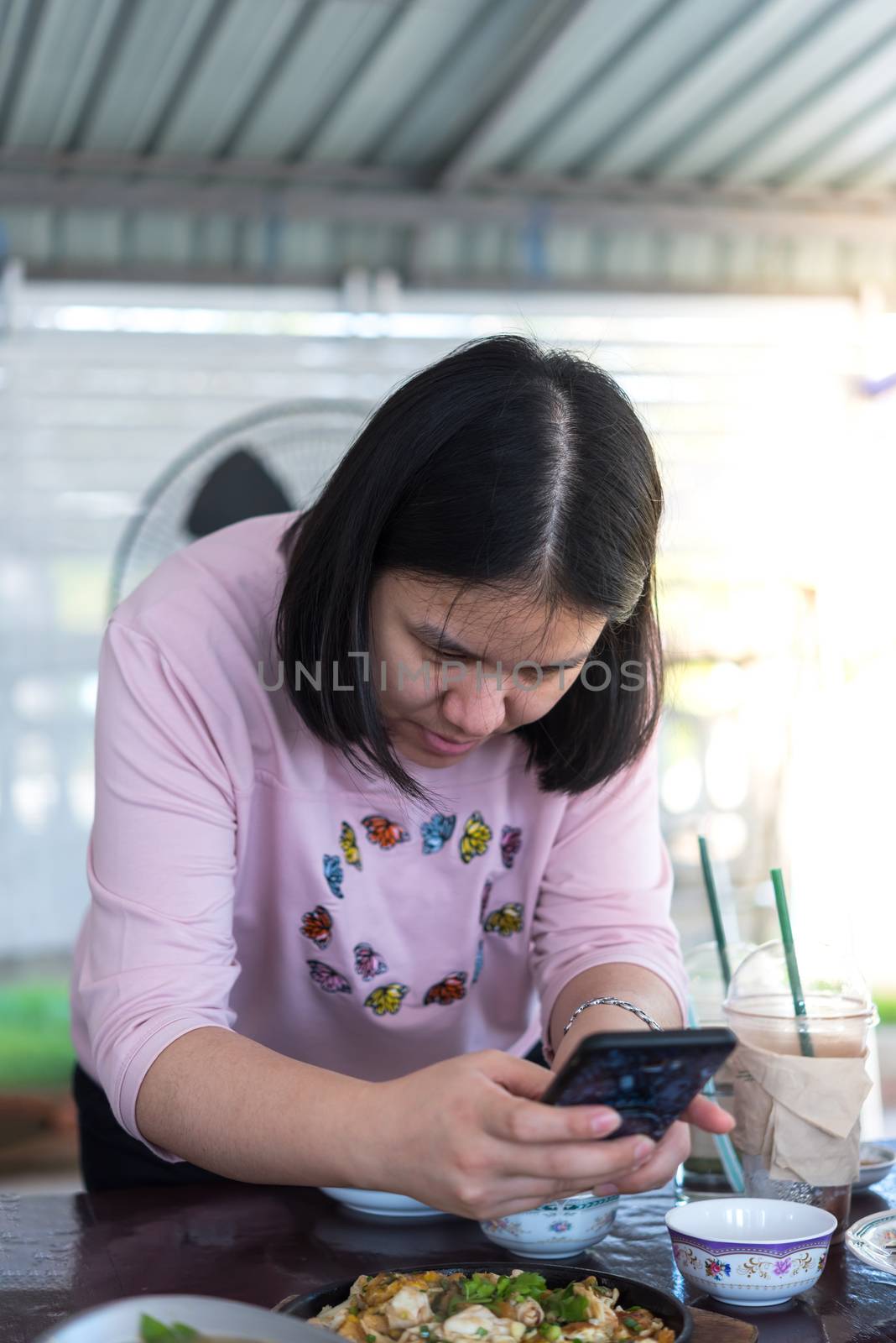 Asian woman foodstagramming use smartphone photographing a food on table in restaurant before eat for post and share on internet social media with fun and happy