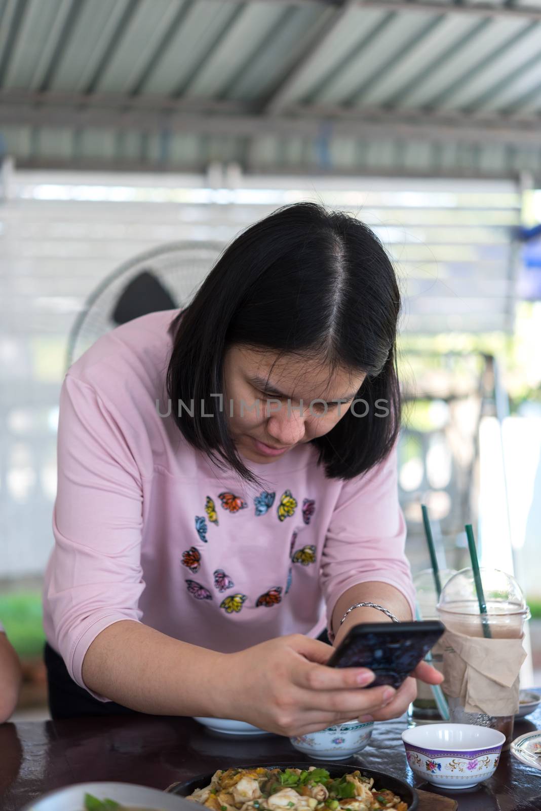 Asian woman foodstagramming use smartphone photographing a food on table in restaurant before eat for post and share on internet social media with fun and happy