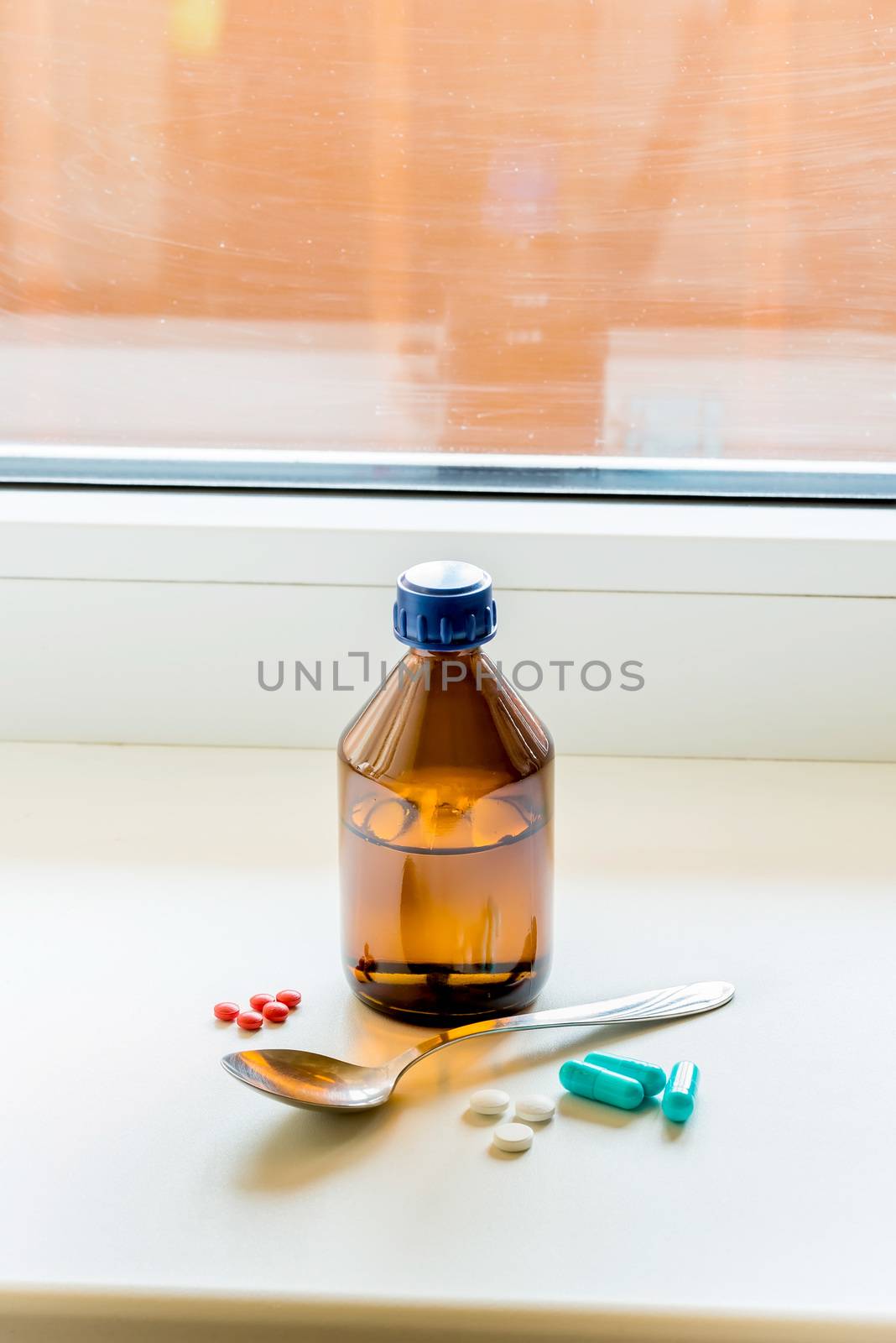 A cough syrup bottle with a spoon and some pills on the table close to the window