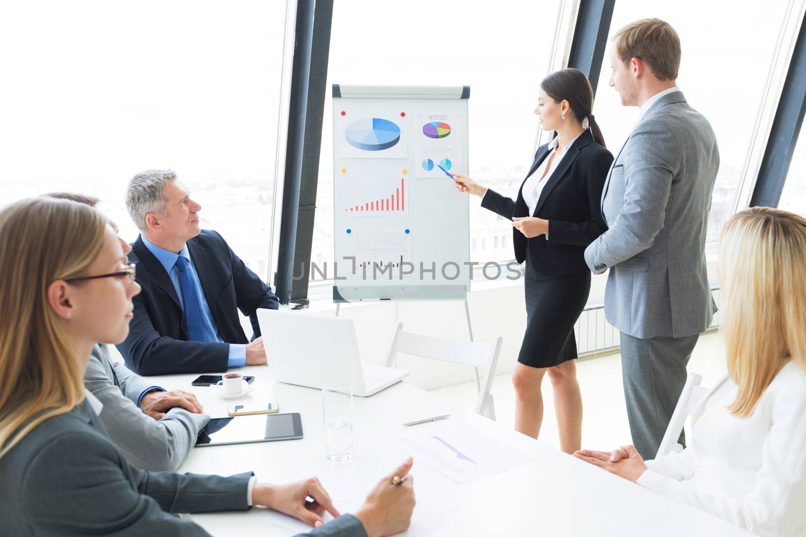 Businesswoman giving presentation using financial diagrams at flipchart to colleagues in office at meeting