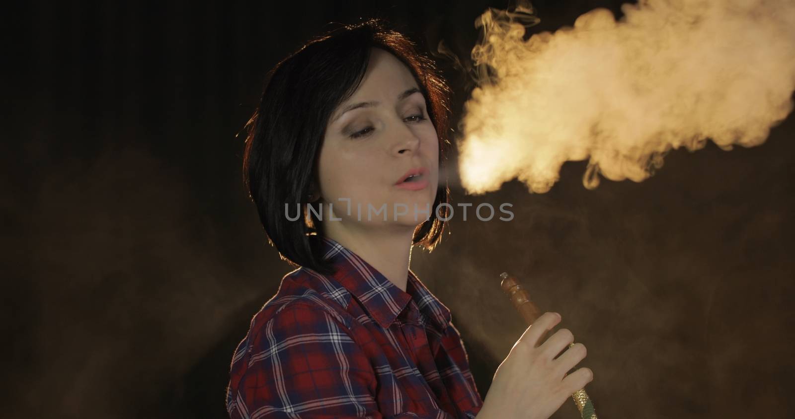 Beautiful, young woman smoking hookah. Attractive girl smoking flavored tobacco. Blow out smoke on black background