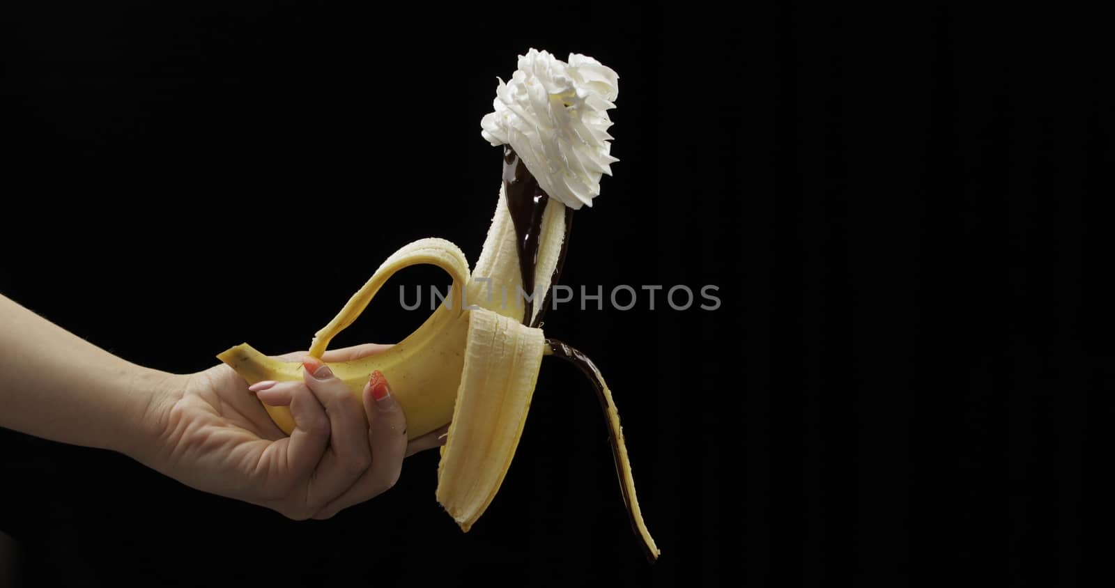 Woman hand holds banana with melted dark chocolate syrup and whipped cream on top of the fruit. A peeled banana covered in whipped cream. Black background