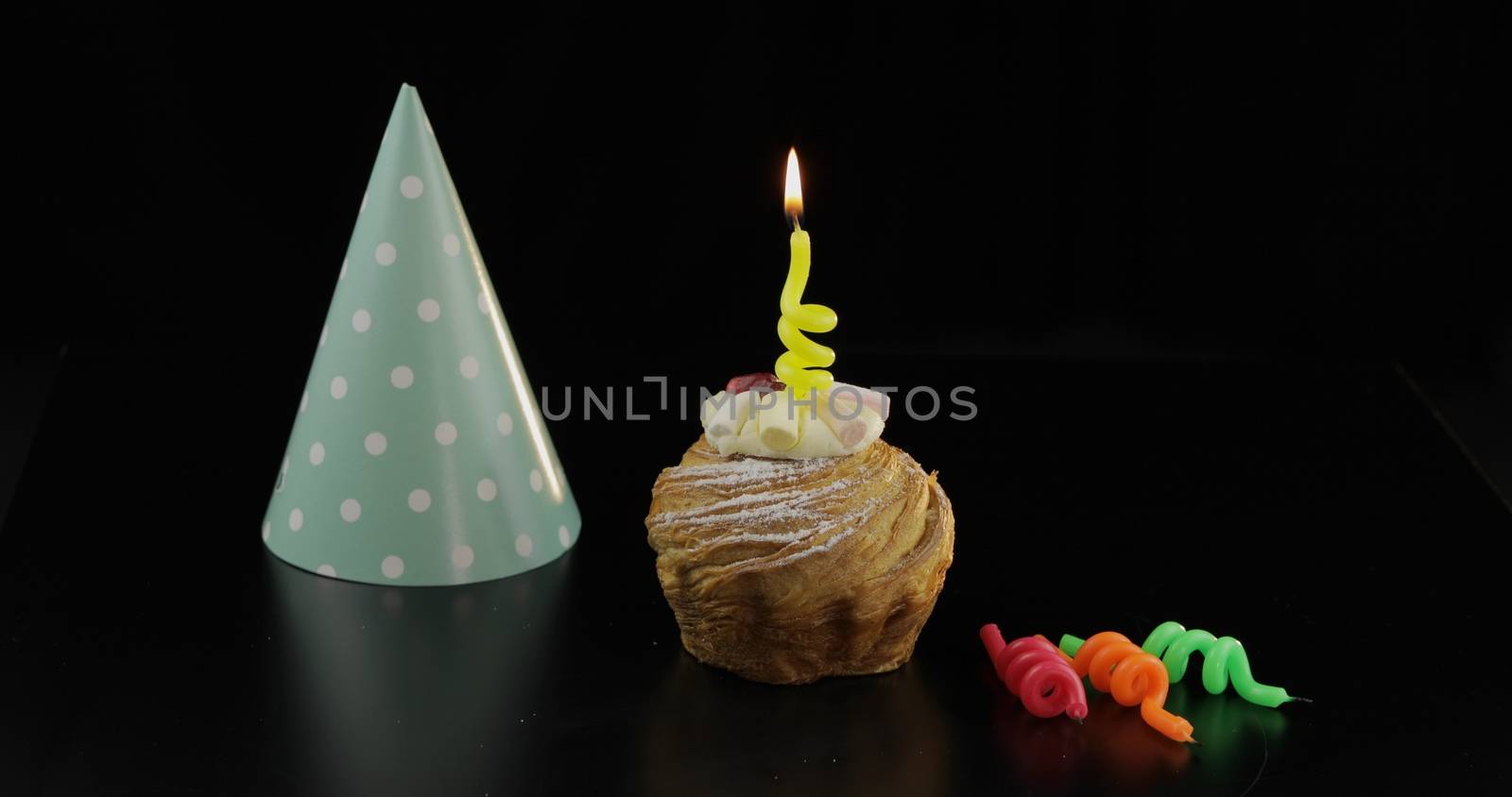 Party. Cake and a yellow festive candle on it. Colored party hat. Black background. Celebrate an event, birthday