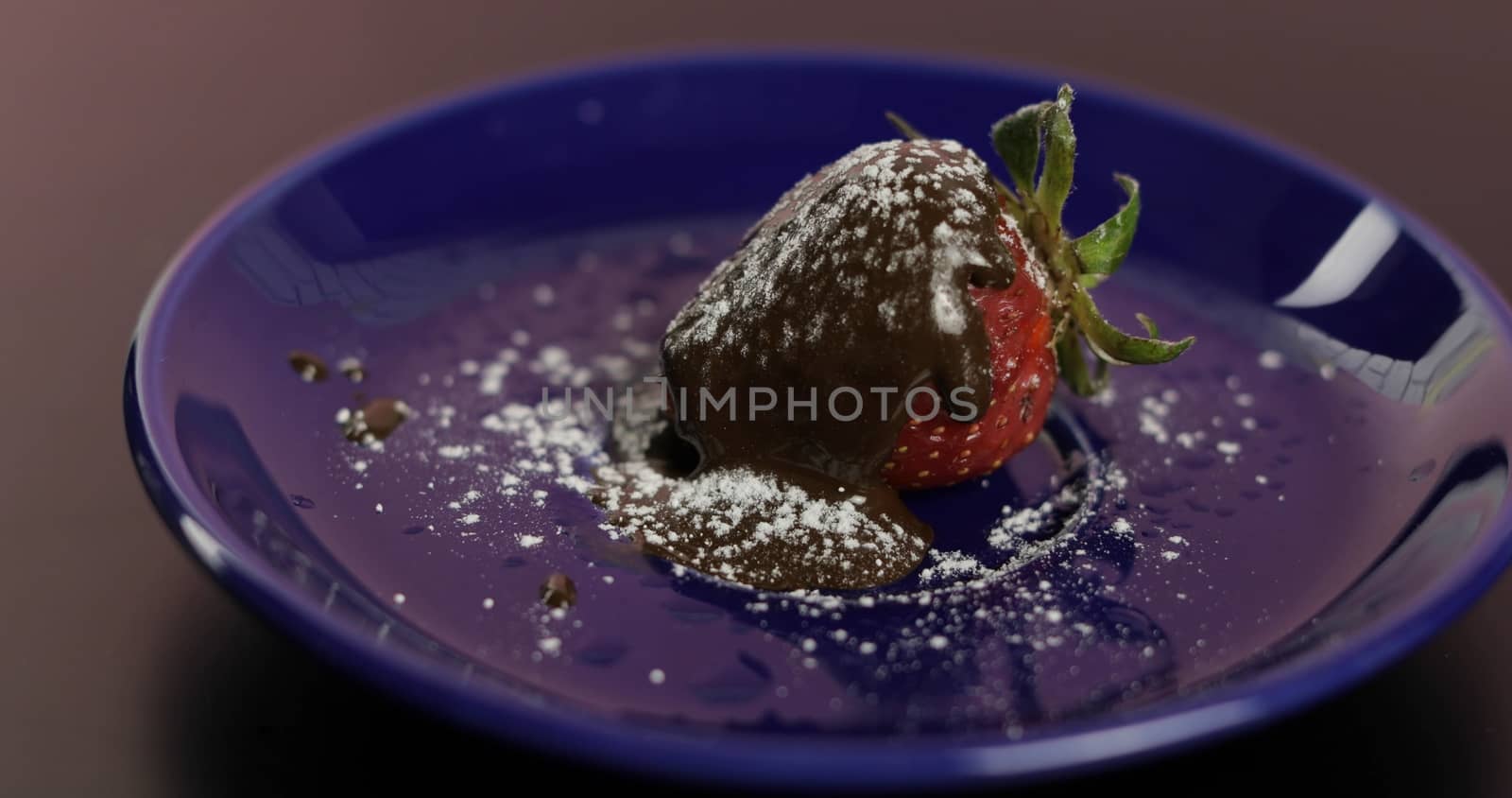 Ripe juicy strawberry in melted dark chocolate with icing sugar powder on a blue plate. Front view. Strawberry in chocolate on a dark background. Close up