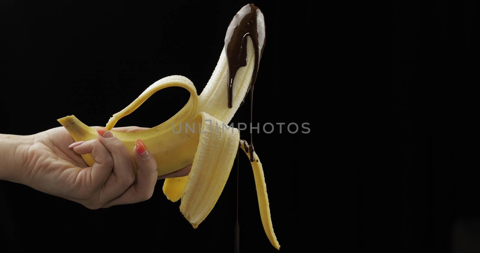 Banana with melted dark chocolate syrup. A peeled banana gets covered in chocolate cream. Black background