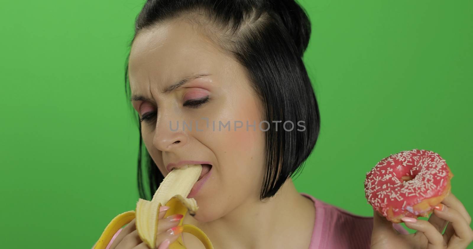 Starting healthy eating. Say no to junk food. Beautiful sad young girl on a chroma key background holds donut in one hand and banana on other. Cute woman choice donut or banana to eat
