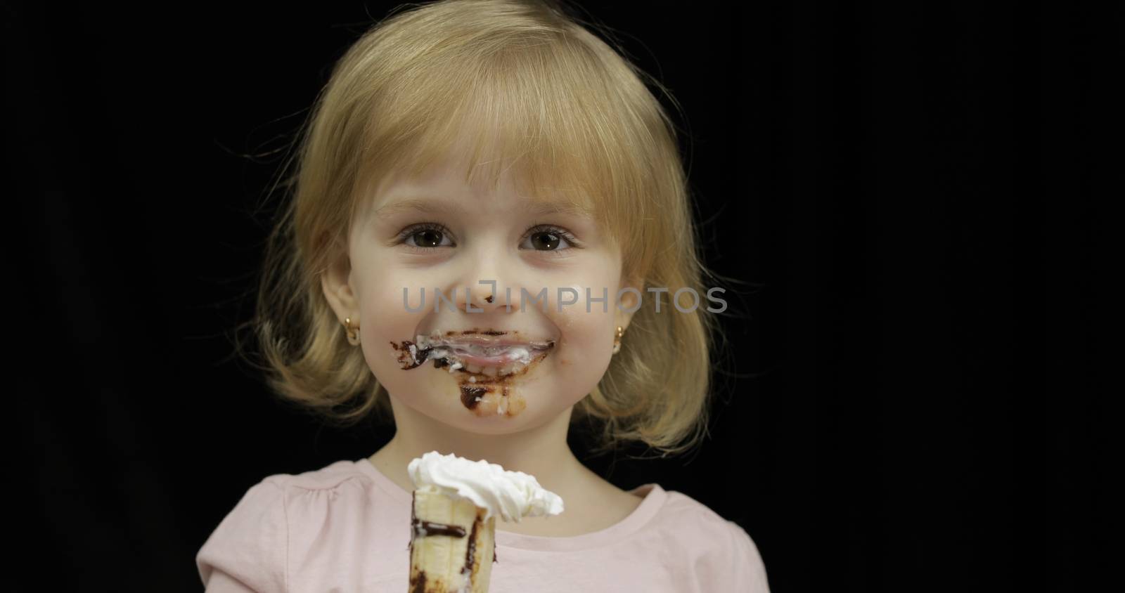 Beautiful, happy child eats banana with melted dark chocolate syrup and whipped cream on black background. Teen blonde girl in pink shirt making faces. Cute smiling. Dirty face