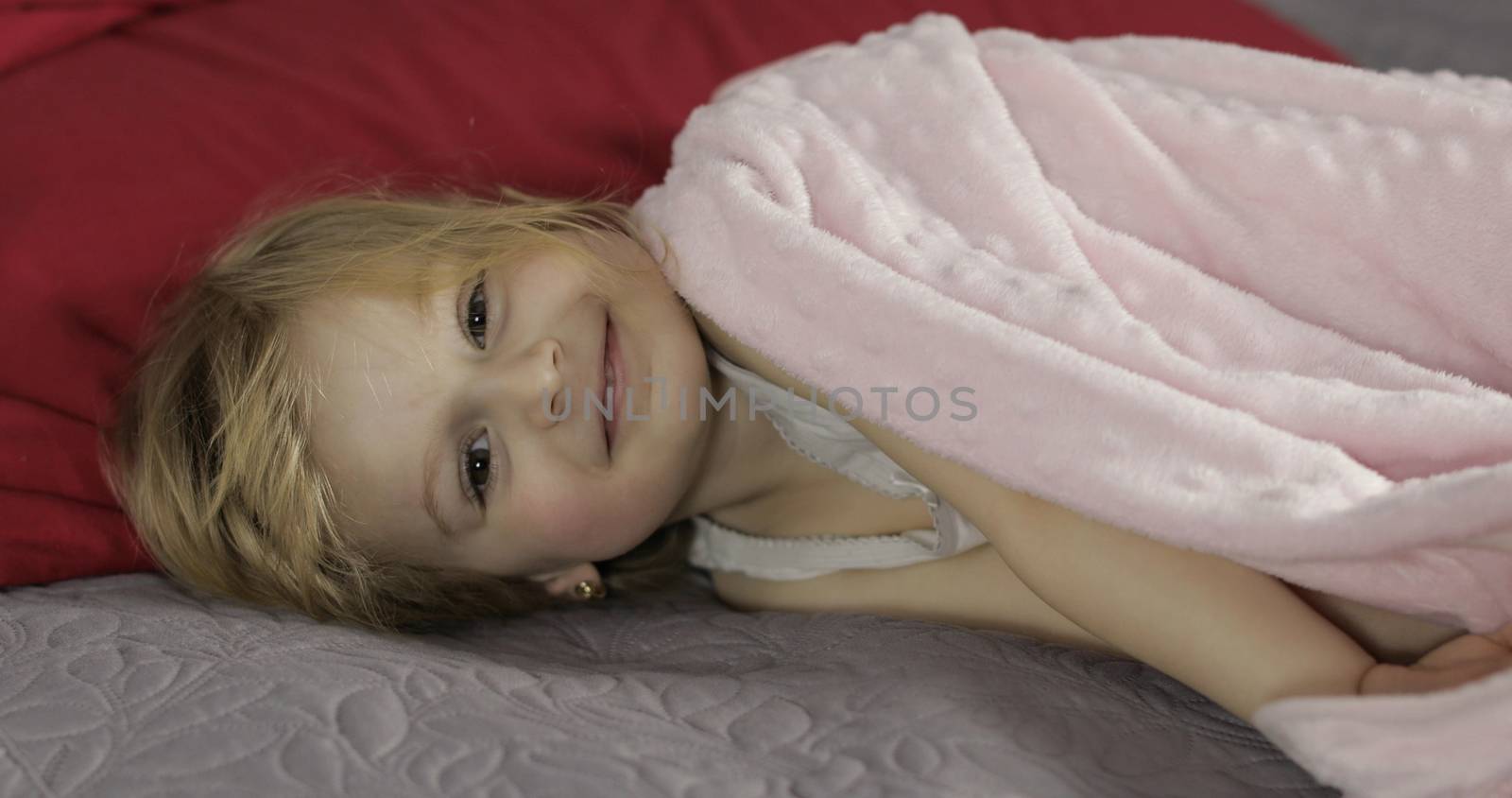 Little cheerful girl under a blanket on cozy bed at home. Pretty, little girl in morning. Sweet, adorable child having fun on the bed under a coverlet. Concept of kids sleep, care or childhood