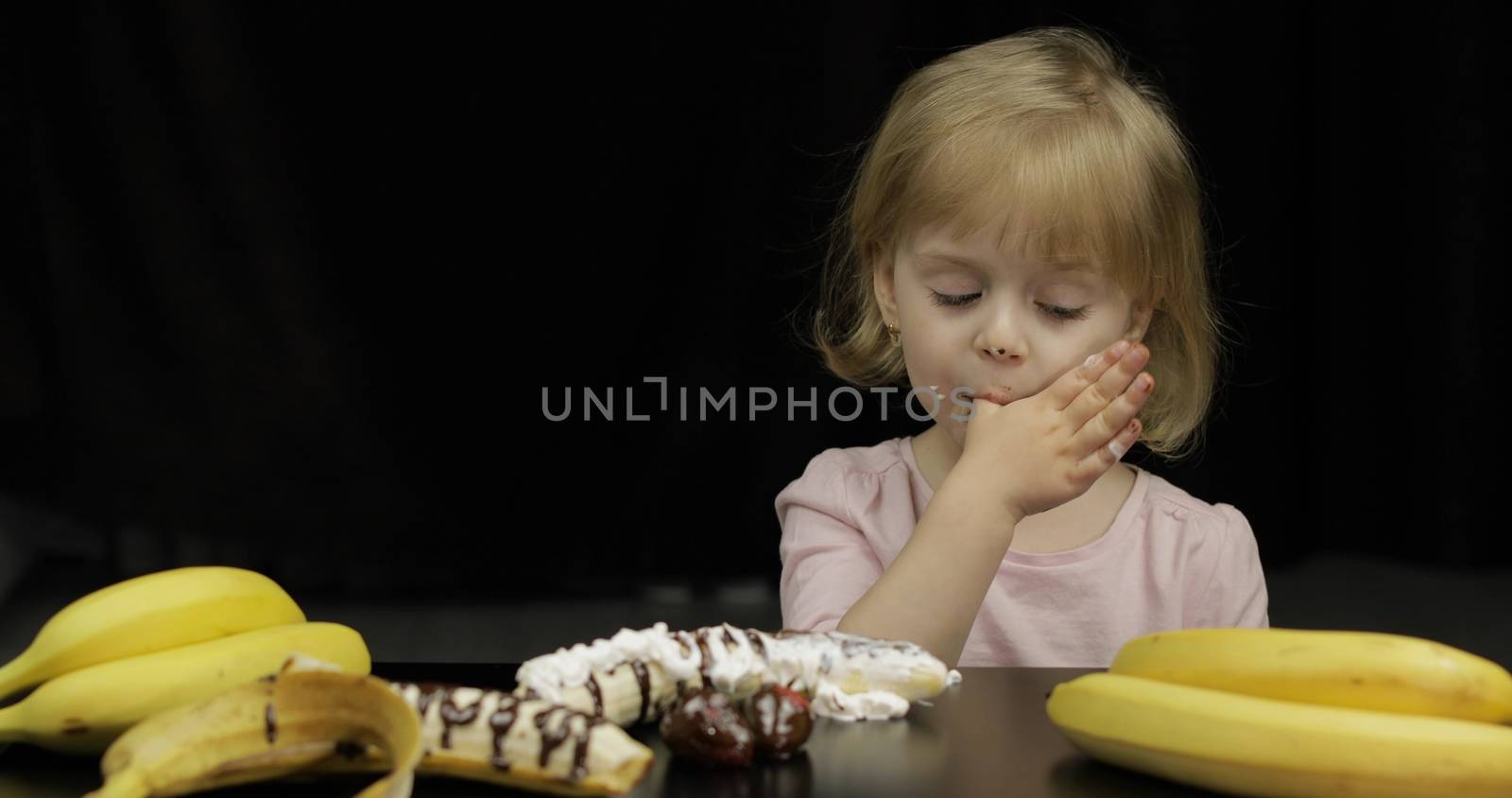 Beautiful, happy child eats melted dark chocolate syrup and whipped cream on black background. Teen blonde girl in pink shirt. Cute smiling. Dirty face