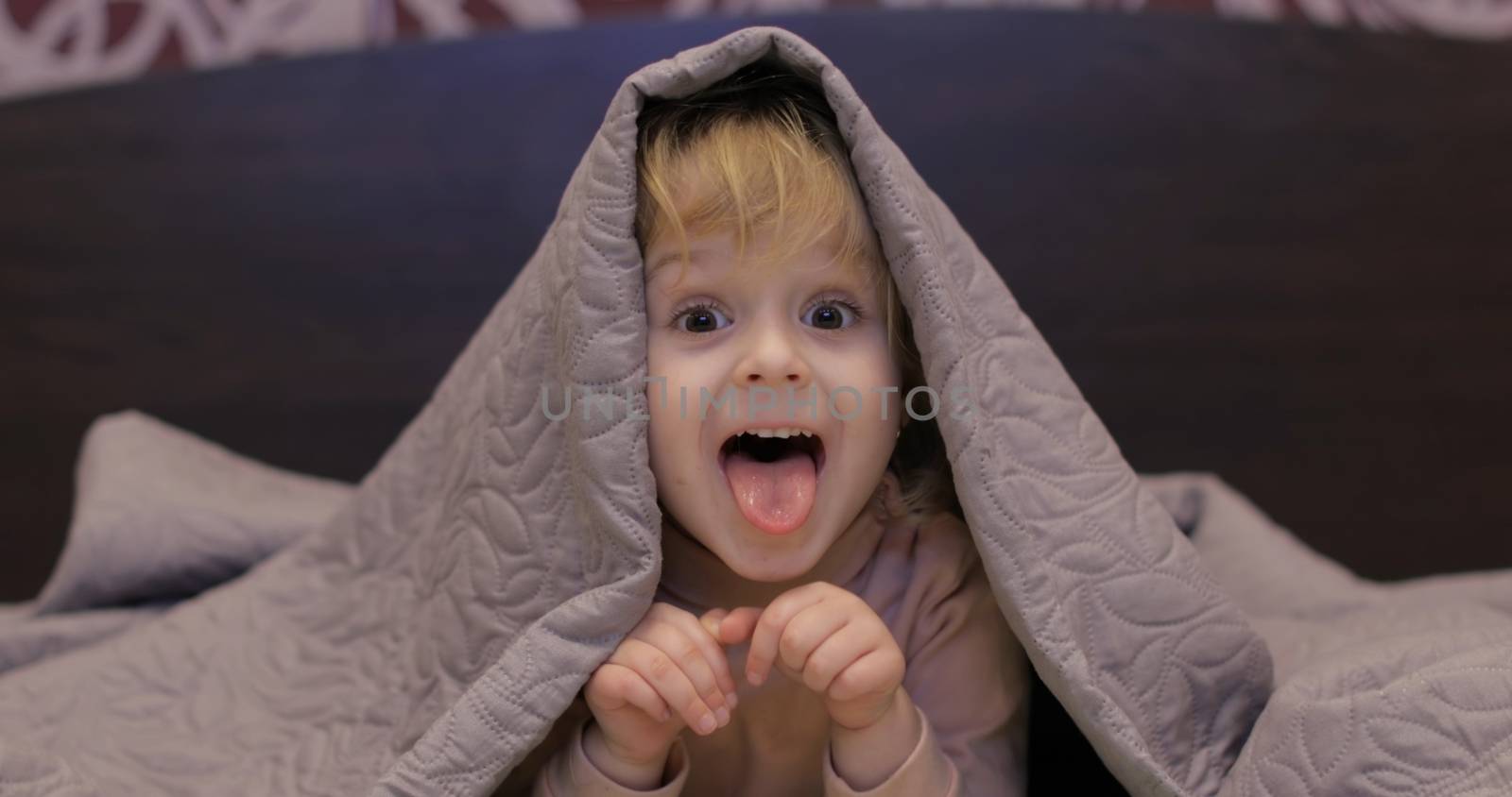 Little cheerful girl hides under a blanket and watching TV. Sweet, adorable child having fun on the bed under a coverlet. Concept of kids sleep, care or childhood