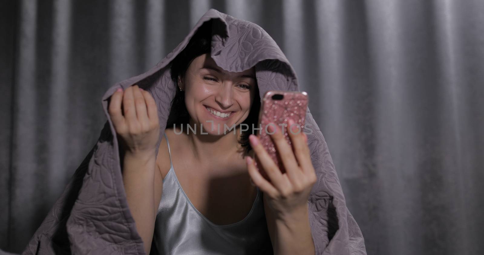 Pretty young brunette woman sitting on the bed under a blanket and having video chat using smartphone enjoying chatting to friend. Blue nightgown. Online communication