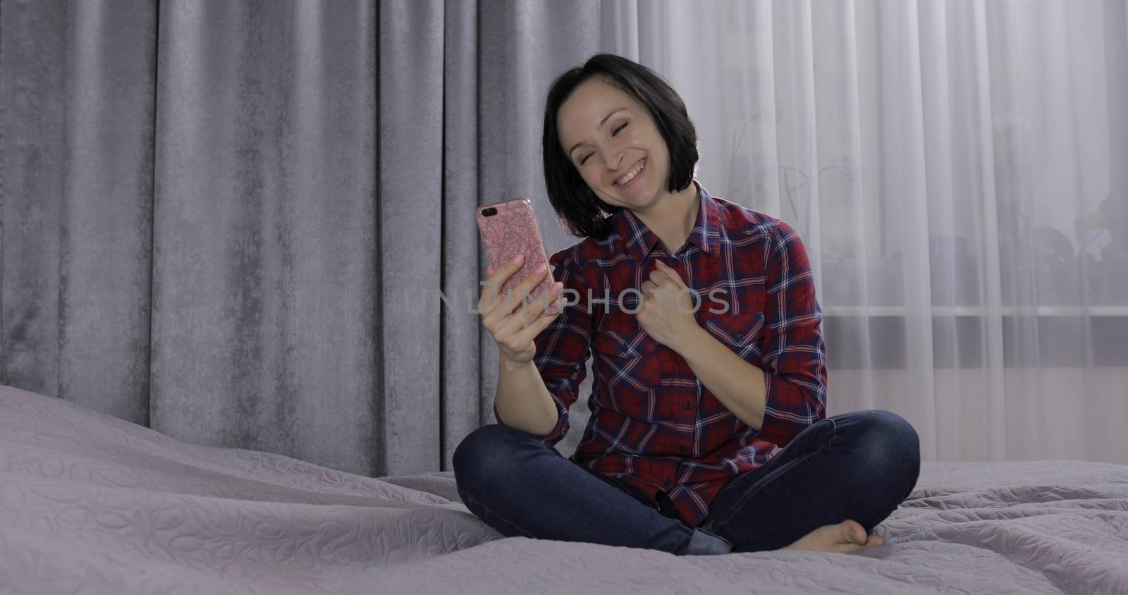 Pretty young brunette woman sit on the bed and having video chat using smartphone enjoying chatting to friend. Indoors. Red shirt and jeans. Online communication