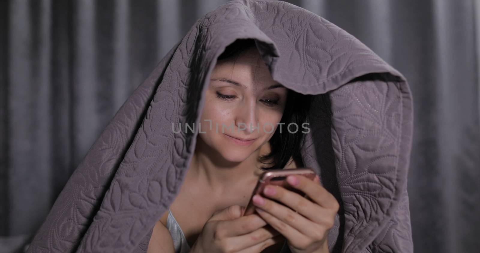 Pretty young brunette woman sitting on the bed under a blanket and having chat on social media using smartphone enjoying chatting to friend. Indoors. Blue nightgown. Online communication