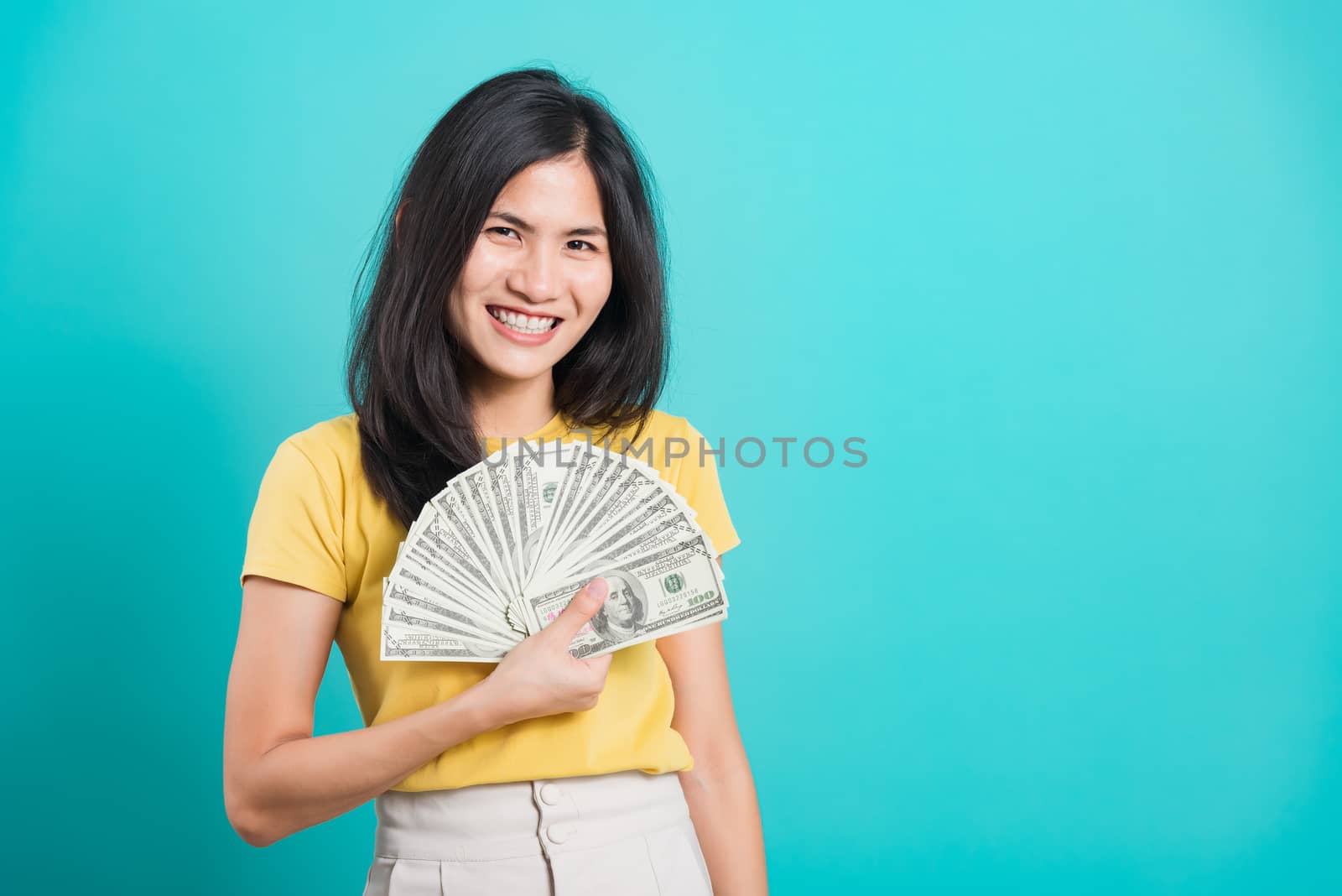 Asian happy portrait beautiful young woman standing wear t-shirt smiling holding money fan banknotes 100 dollar bills and looking to camera isolated on blue background with copy space for text