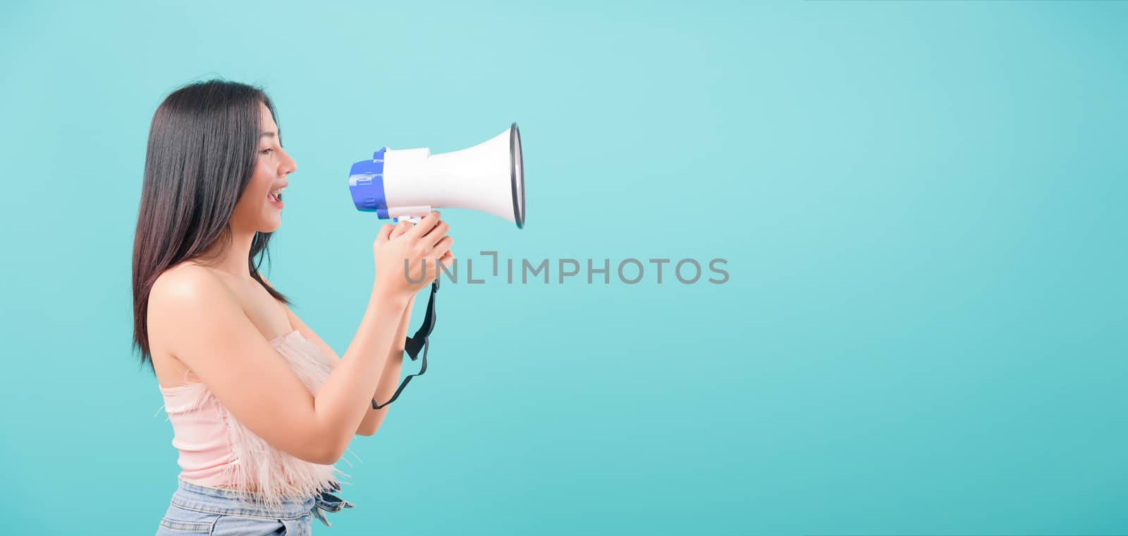 Asian happy portrait beautiful young woman standing smile her  holding her Shouting through a megaphone message to something and looking to camera on blue background with copy space for text