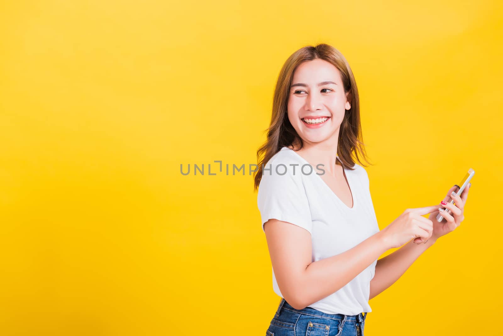 Portrait Asian Thai beautiful happy young woman standing smile, holding mobile phone her looking back to space, studio shot isolated on yellow background, with copyspace