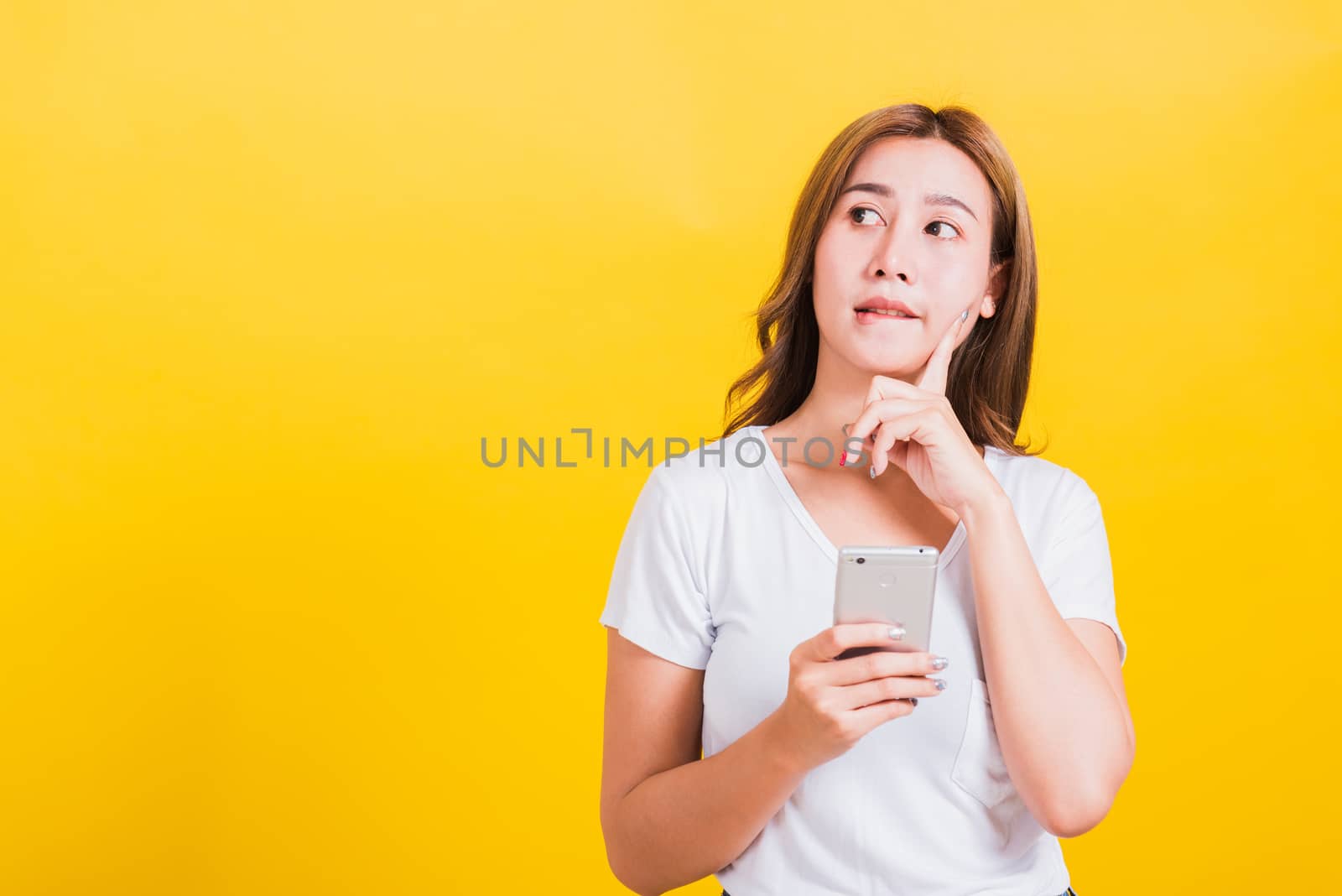 Asian Thai portrait happy beautiful cute young woman stand wear t-shirt her using holding smart mobile phone and chin handle thinking looking to side, studio shot isolated yellow background copy space