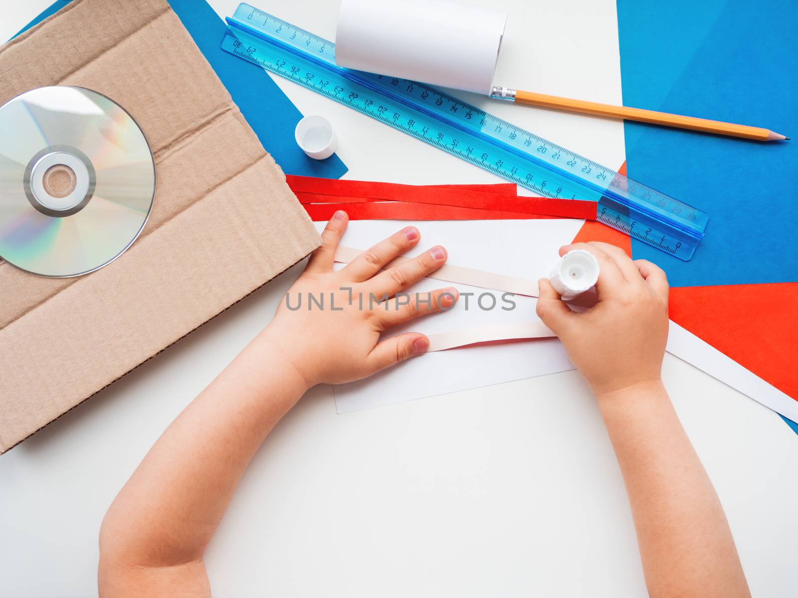 Child makes something out of colored paper, toilet paper core, carton, scissors, glue, CD-disk, pencil and ruler. Red, white and blue pallette. Making toys from improvised materials.