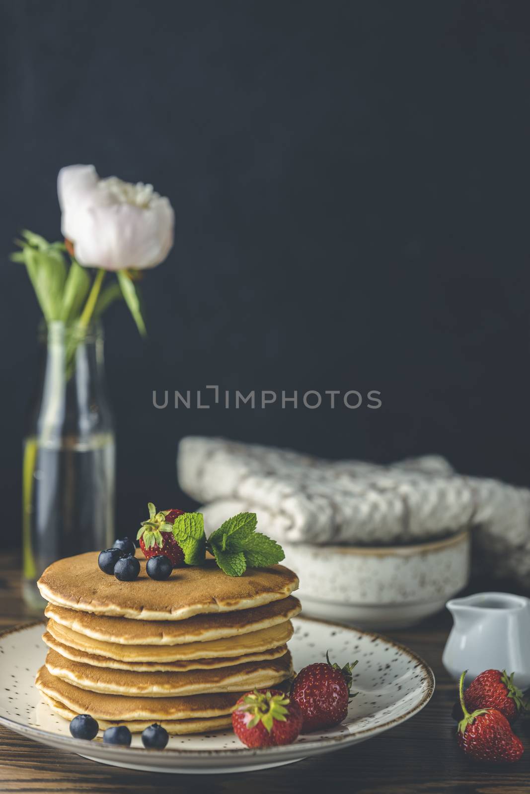 Pancake with strawberry, blueberry and mint in ceramic dish, syrup from small ceramic jar and flowers on a dark wooden table and black background. 