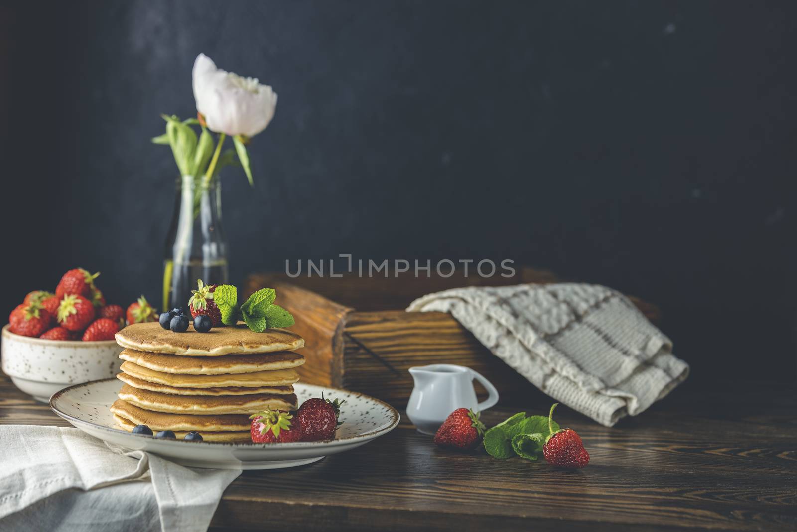 Pancake with srtawberry, blueberry and mint in ceramic dish, syrup from small ceramic jar and flowers on a dark wooden table and black background. 