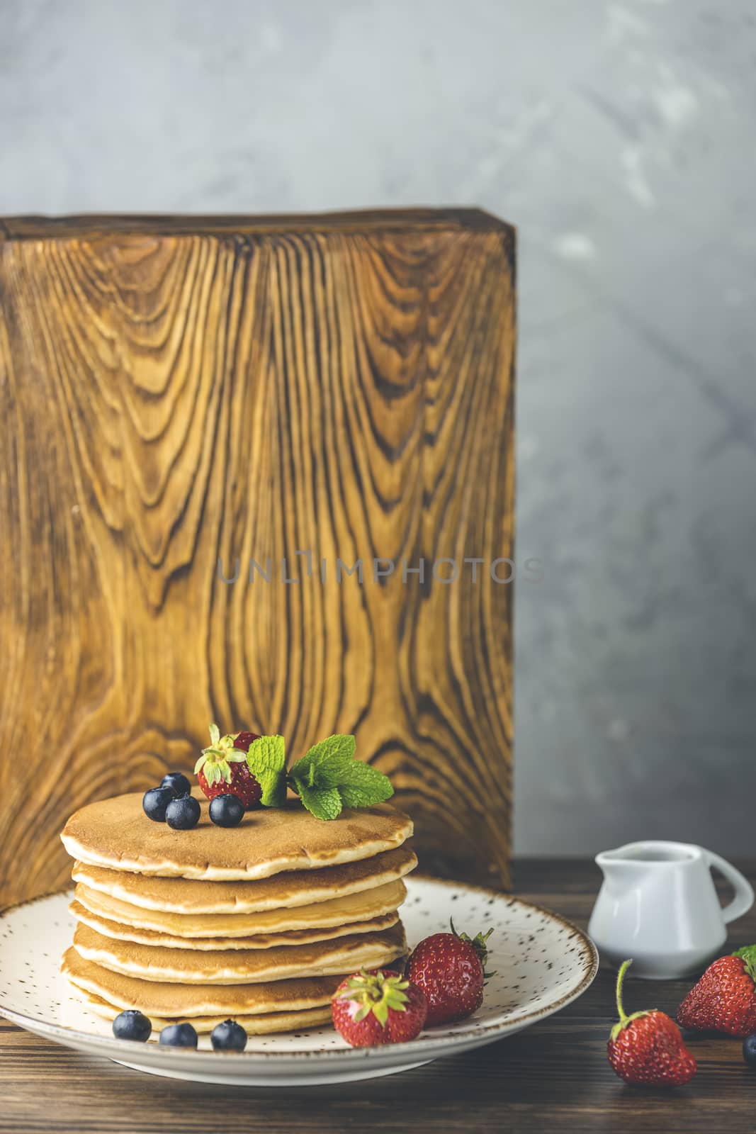 Pancake with srtawberry, blueberry and mint in ceramic dish, syrup from small ceramic jar on a light wooden table and gray background. 