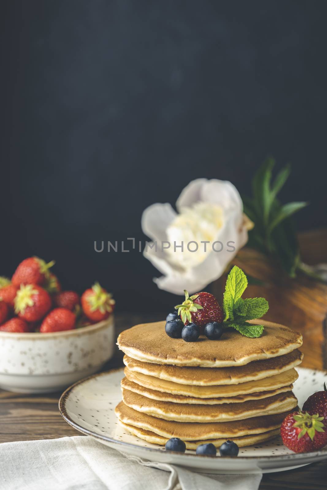 Pancake with srtawberry, blueberry and mint in ceramic dish, syrup from small ceramic jar and flowers on a dark wooden table and black background. 
