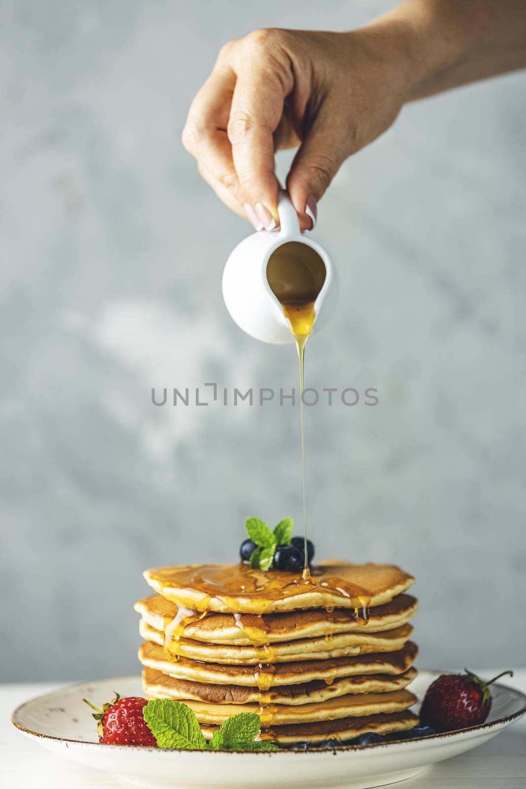 Women hand is pouring syrup from small ceramic jar to pancake with srtawberry, blueberry and mint in ceramic dish on a light wooden table and gray background.