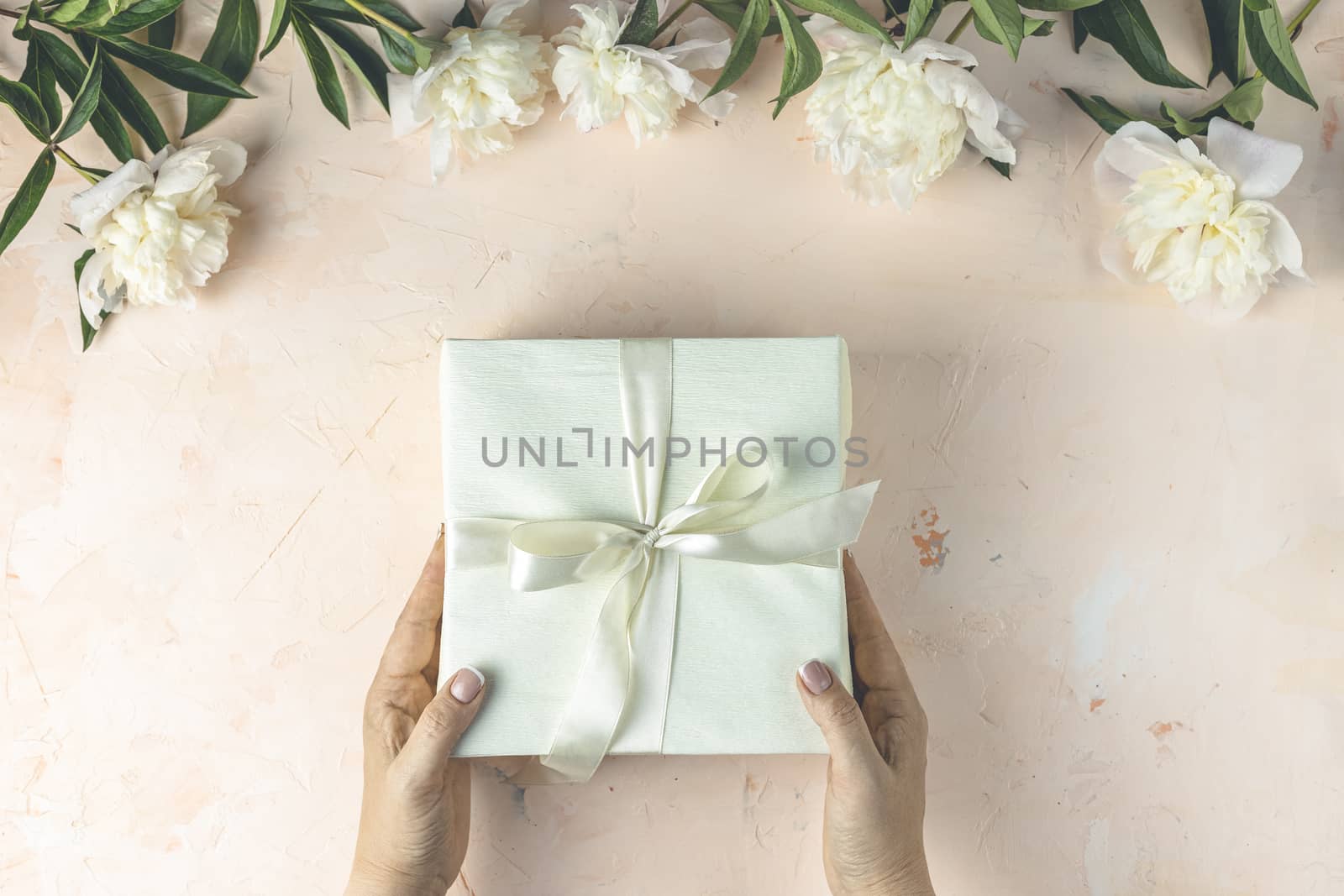 Womans hands hold gift on light concrete table surface surrounded beautiful white peony flowers. Top view, flat lay, greeting card. Feminine flat lay composition with flowers and present.