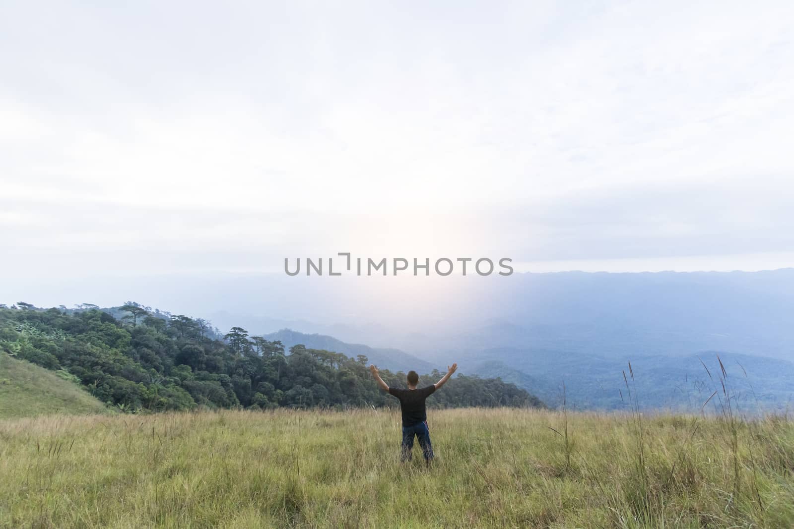 Happy young man standing with raised arms in green field on mountains