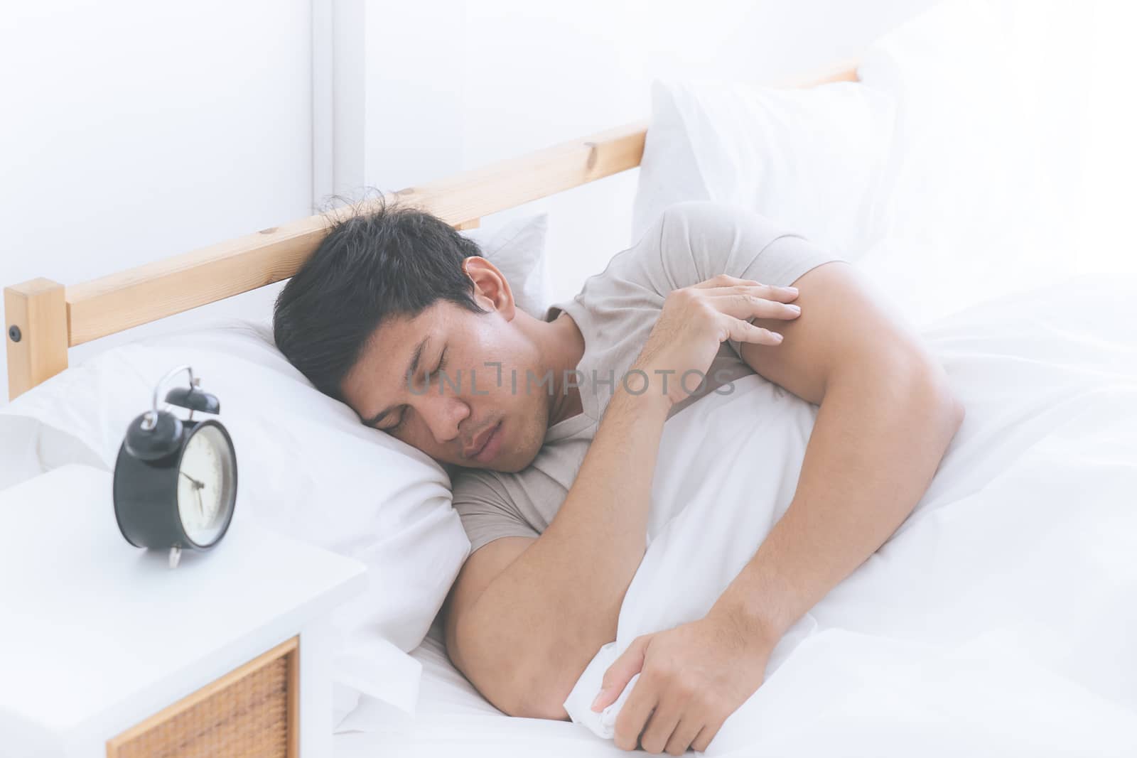 Asian man is sleeping side way on white bed with alarm clock by junce