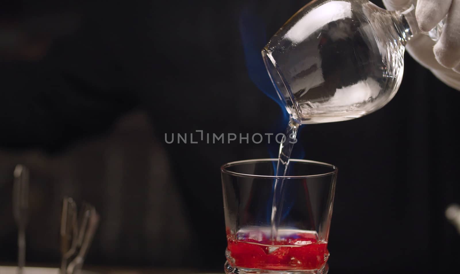 Making of Raspberry Sambuca. Close up barmans hand sets fire to sambuca in snifter and pours it into a glass with raspberries.