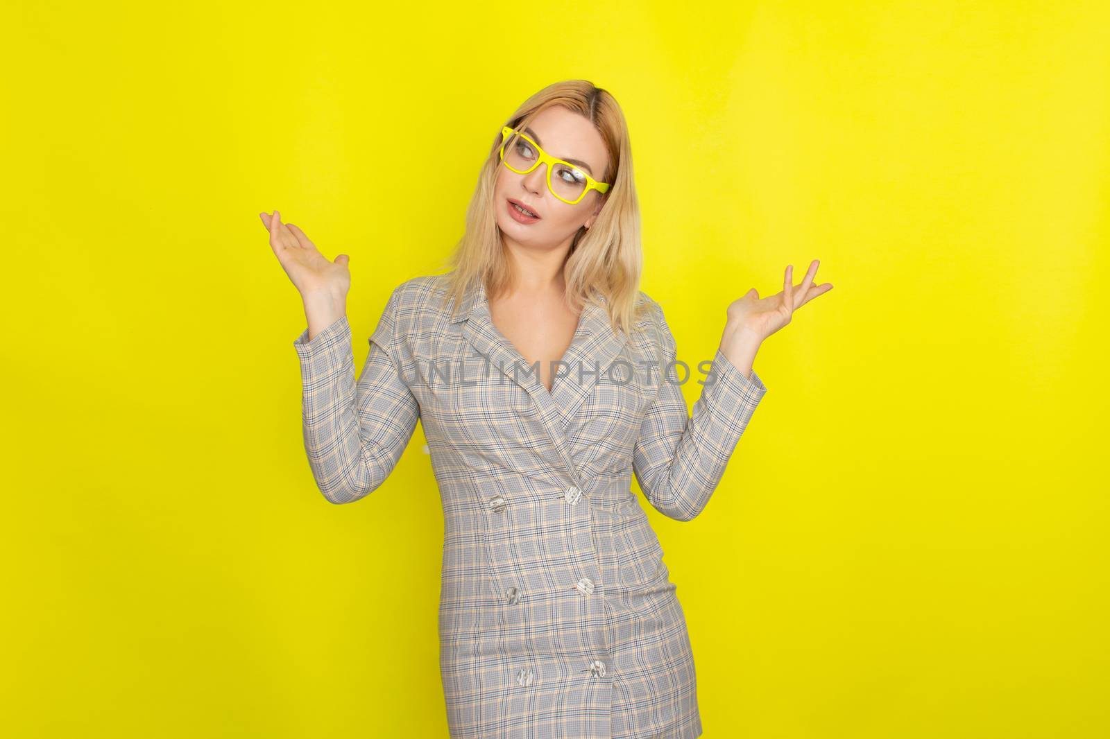 Attractive blonde woman in plaid jacket dress over yellow background wearing yellow glasses