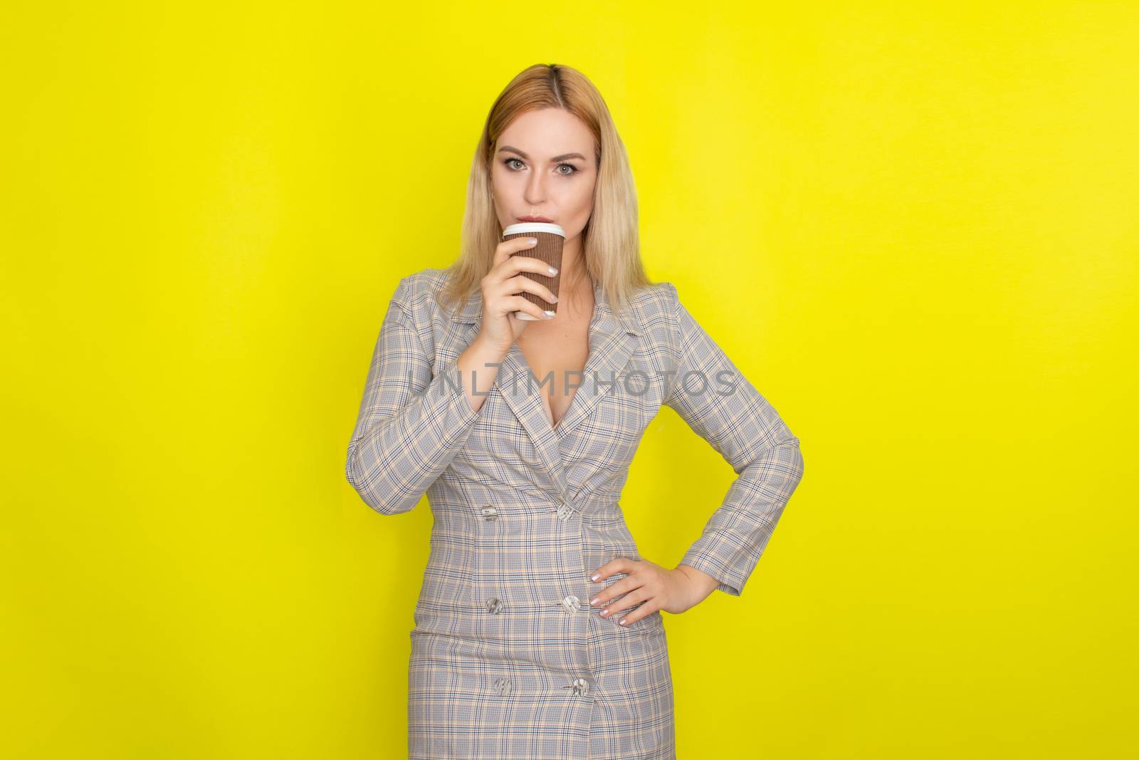 Business blonde woman wearing plaid jacket style dress and drinking coffee over yellow background