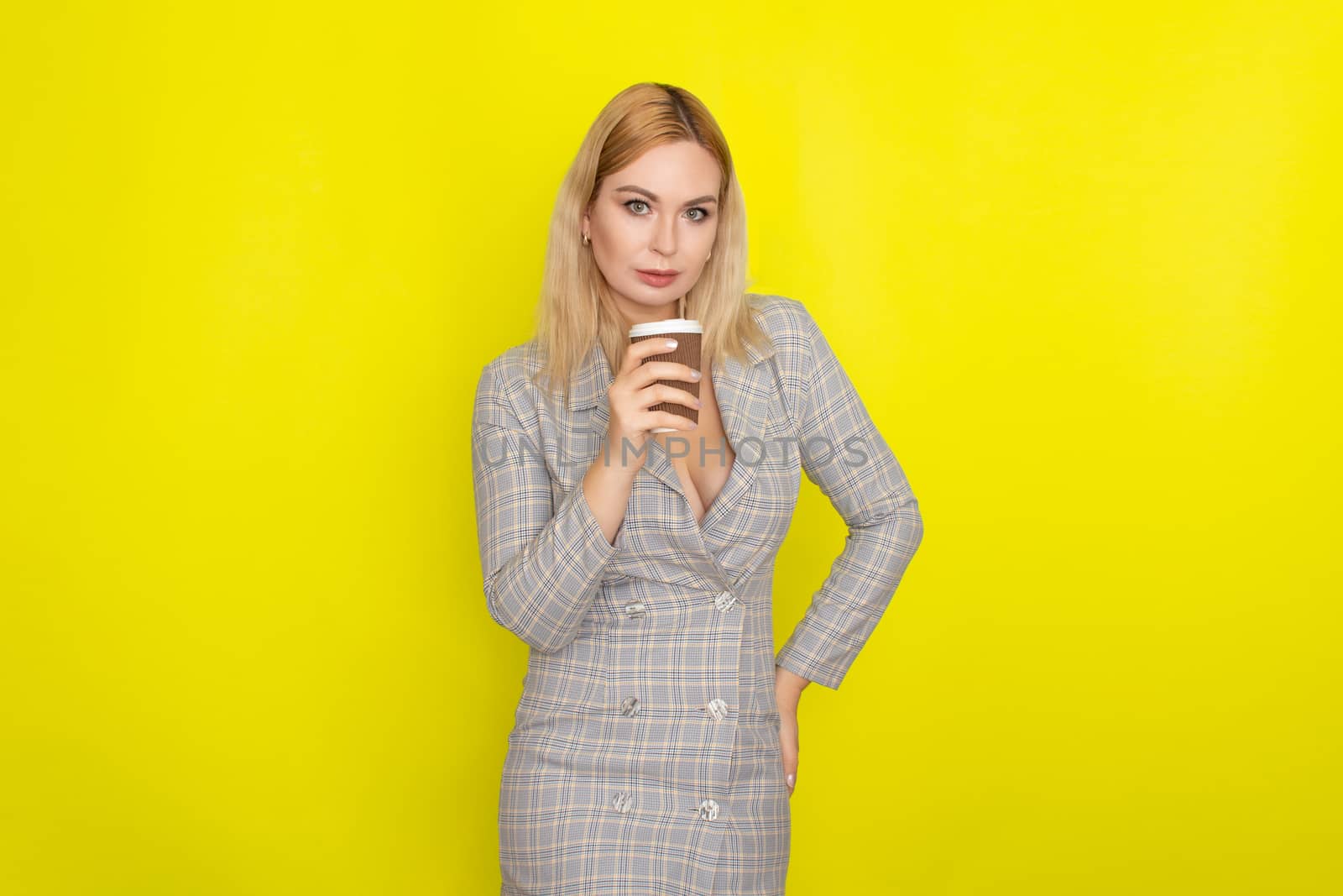 Business blonde woman wearing plaid jacket style dress and drinking coffee over yellow background