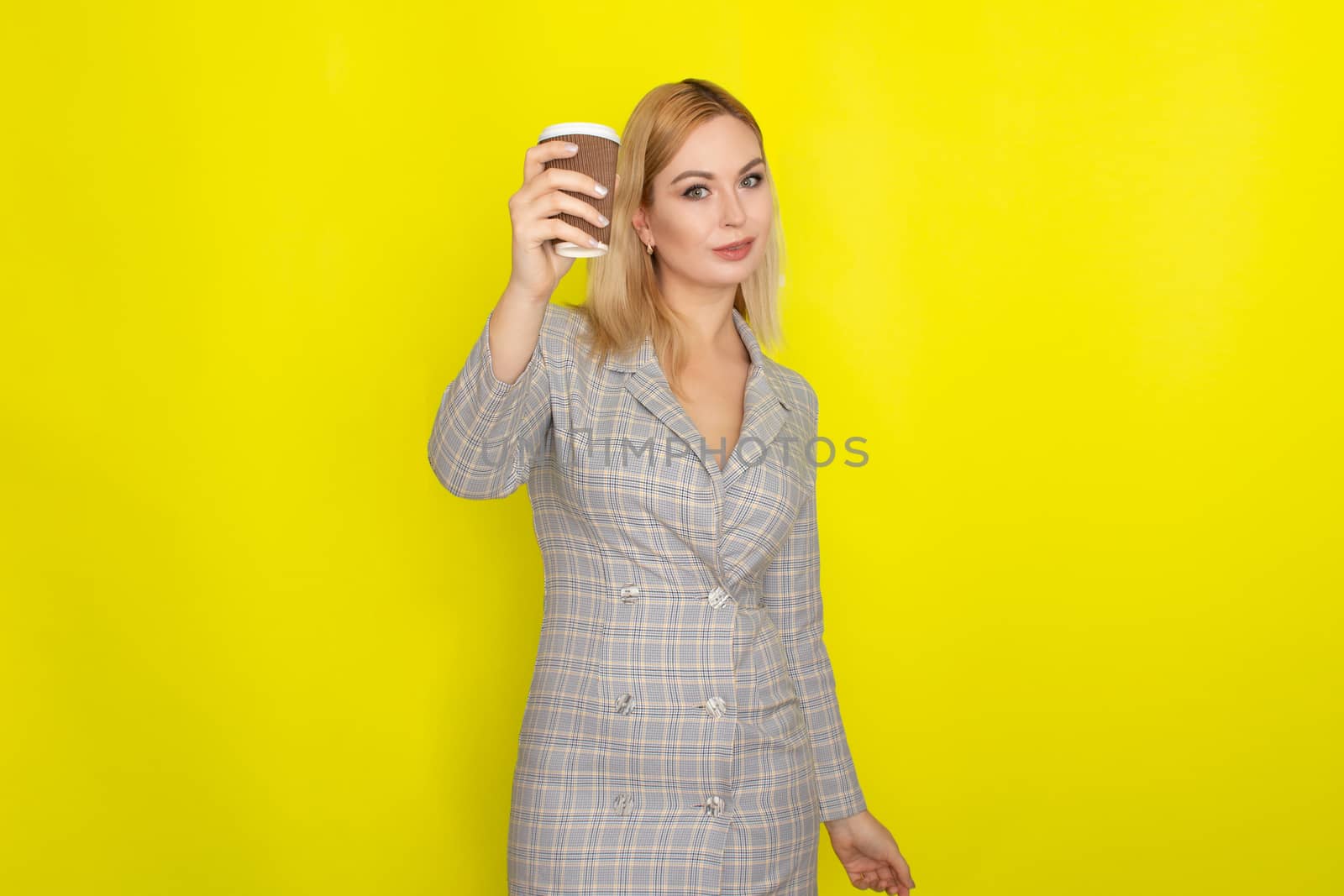 Business blonde woman wearing plaid jacket style dress and drinking coffee over yellow background