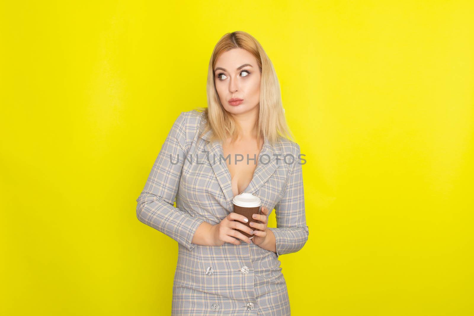 Business blonde woman wearing plaid jacket style dress and drinking coffee over yellow background