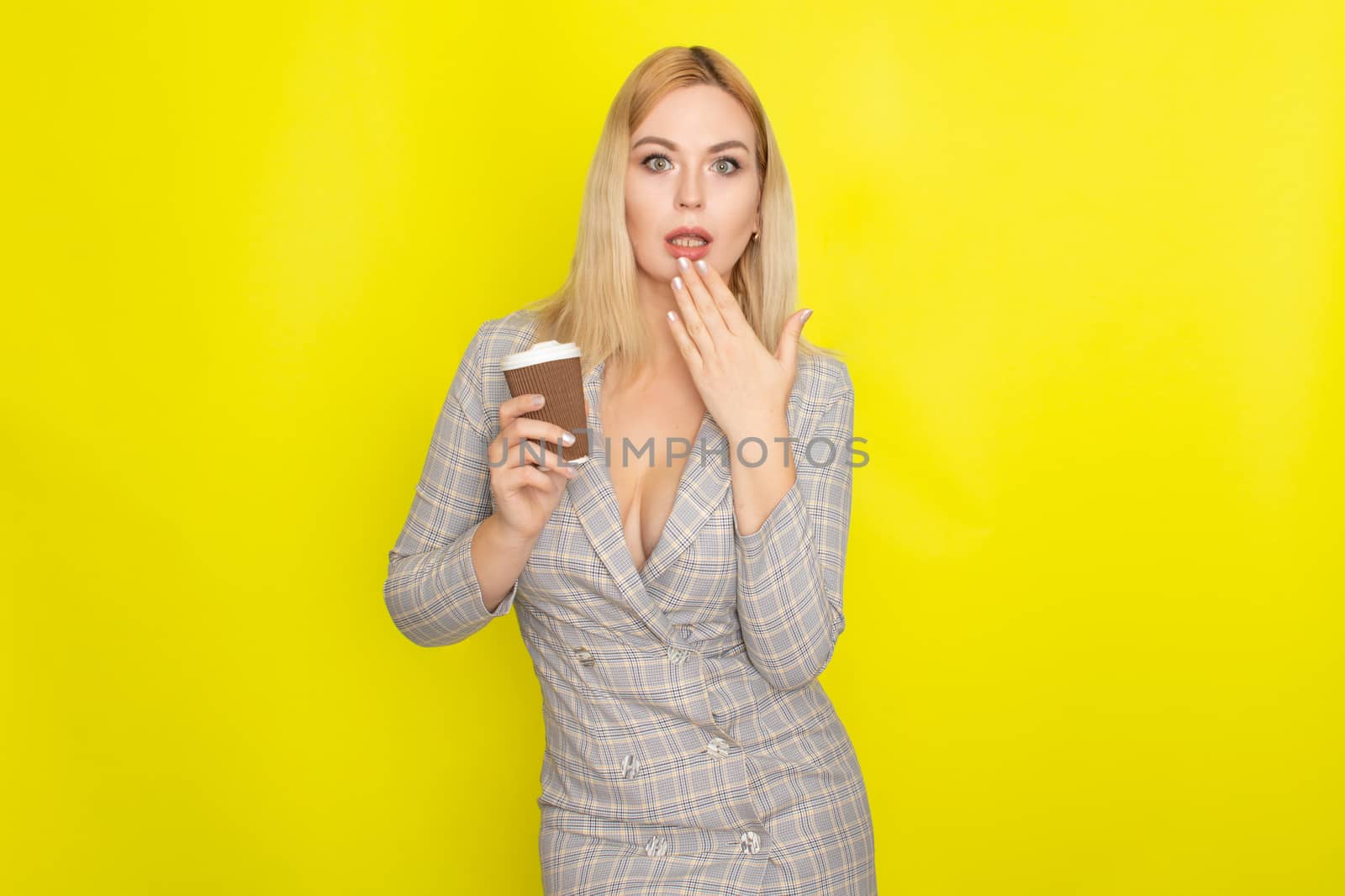 Business blonde woman wearing plaid jacket style dress and drinking coffee over yellow background