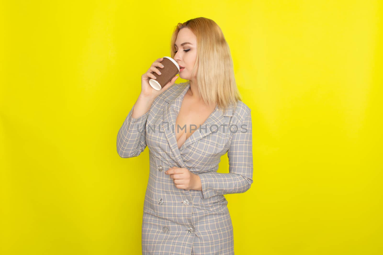 Business blonde woman wearing plaid jacket style dress and drinking coffee over yellow background