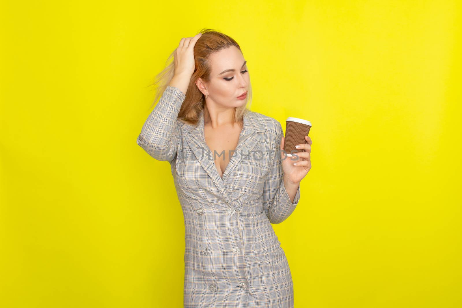 Business blonde woman wearing plaid jacket style dress and drinking coffee over yellow background