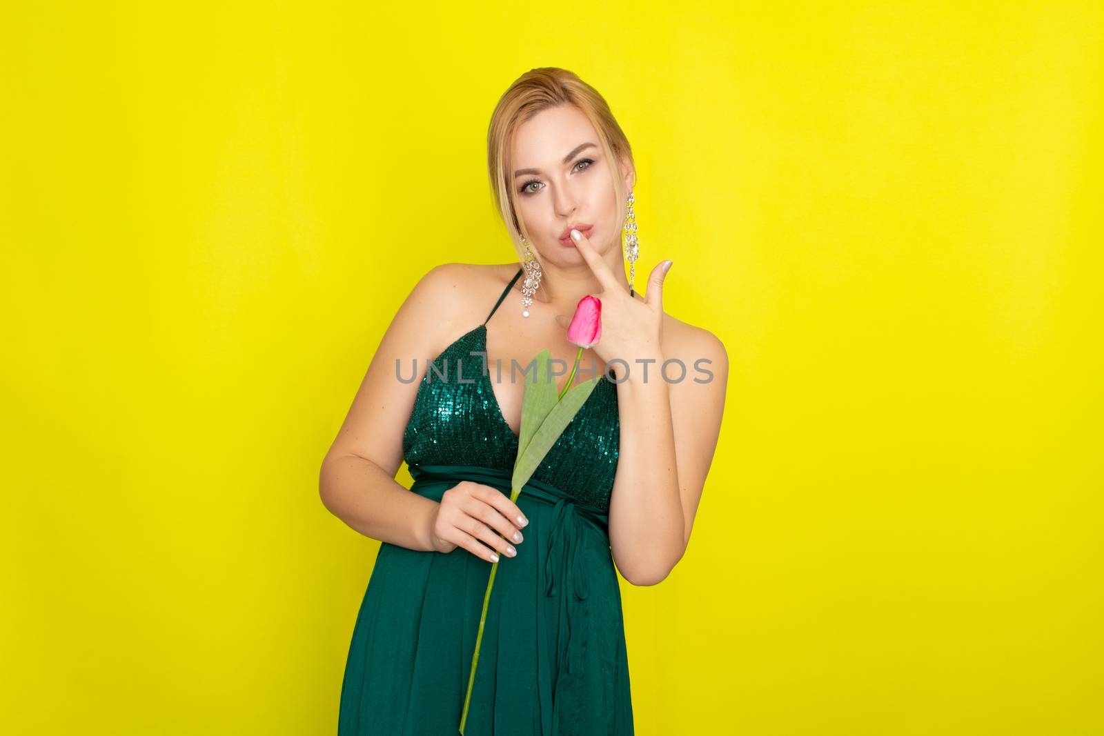 Blonde woman in green evening dress holding one tulip in her hands over yellow background