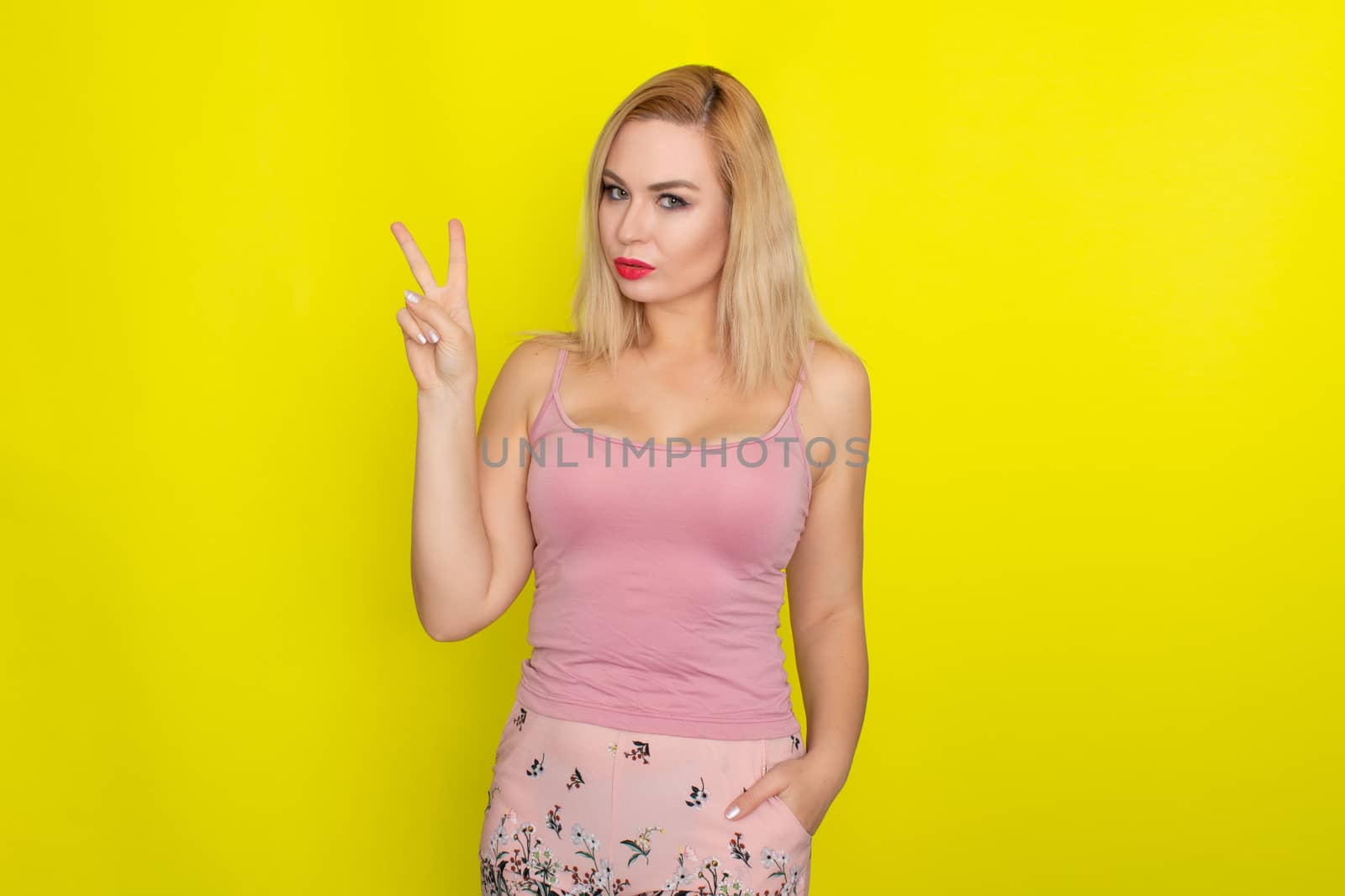 Indoor summer closeup portrait of young stylish fashion glamorous blonde woman posing in pink shorts and shirt, standing over yellow background