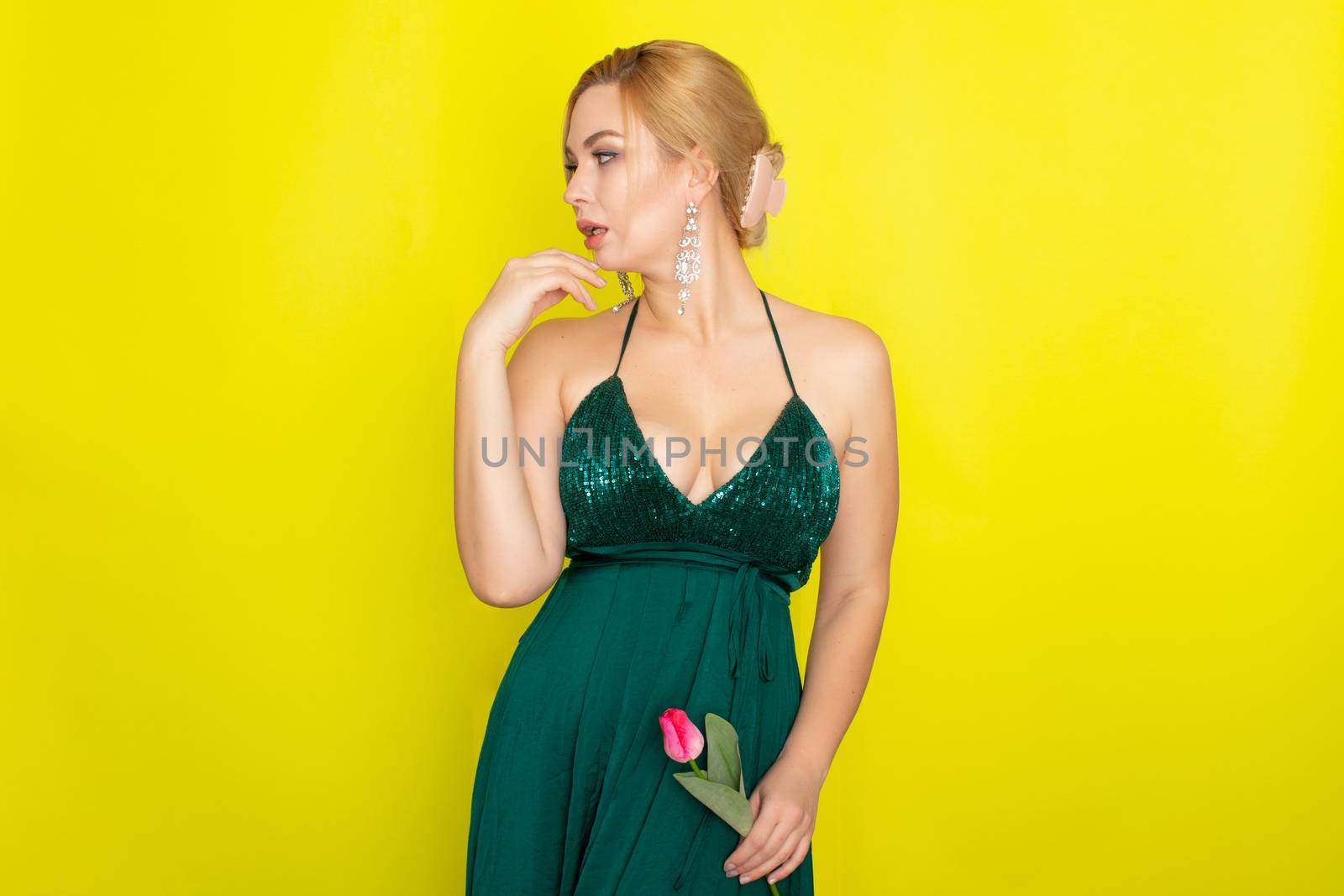 Blonde woman in green evening dress holding one tulip in her hands over yellow background